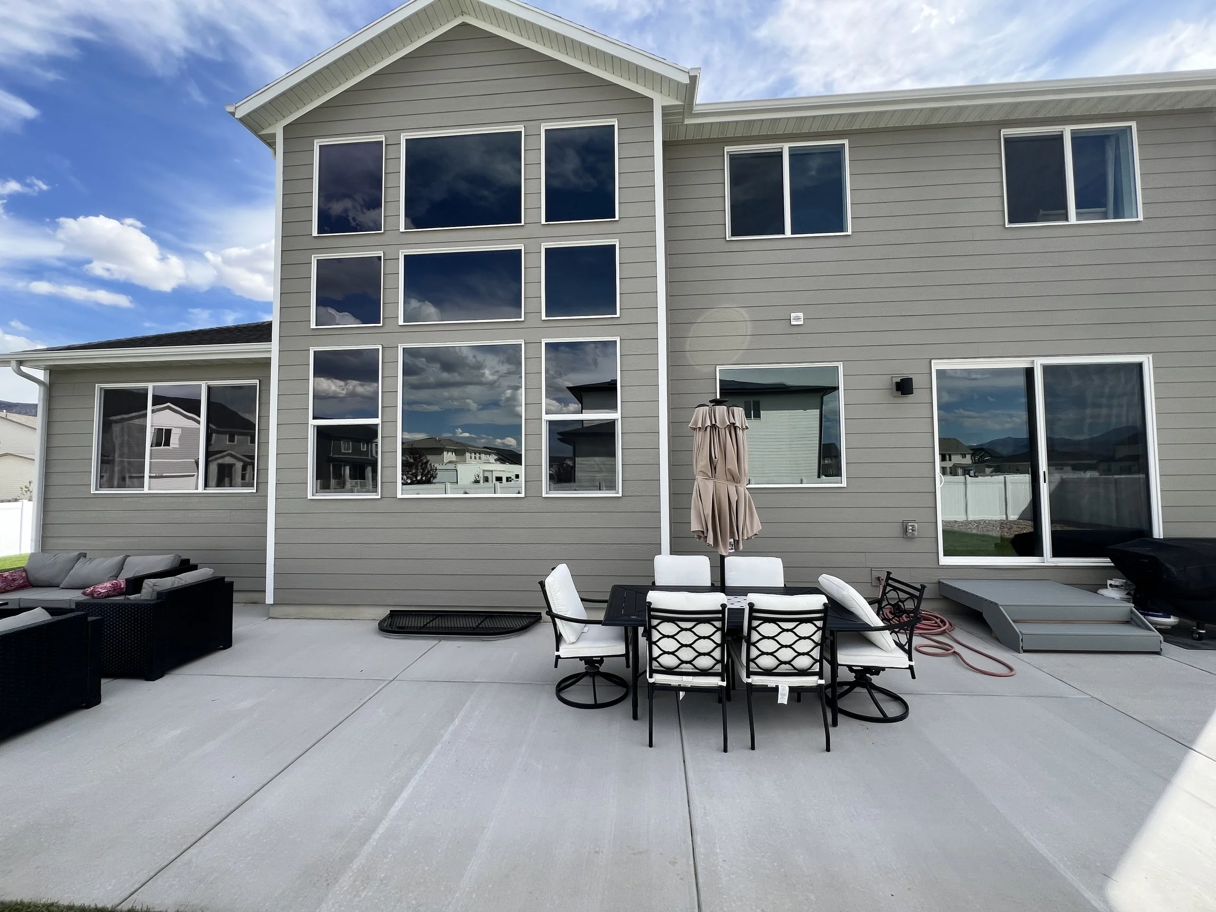 Backyard patio with outdoor furniture including a dining table with six chairs, a sofa set with cushions, and a closed umbrella in front of a modern house with large windows. The house has gray siding and sliding glass doors, under a partly cloudy sky.