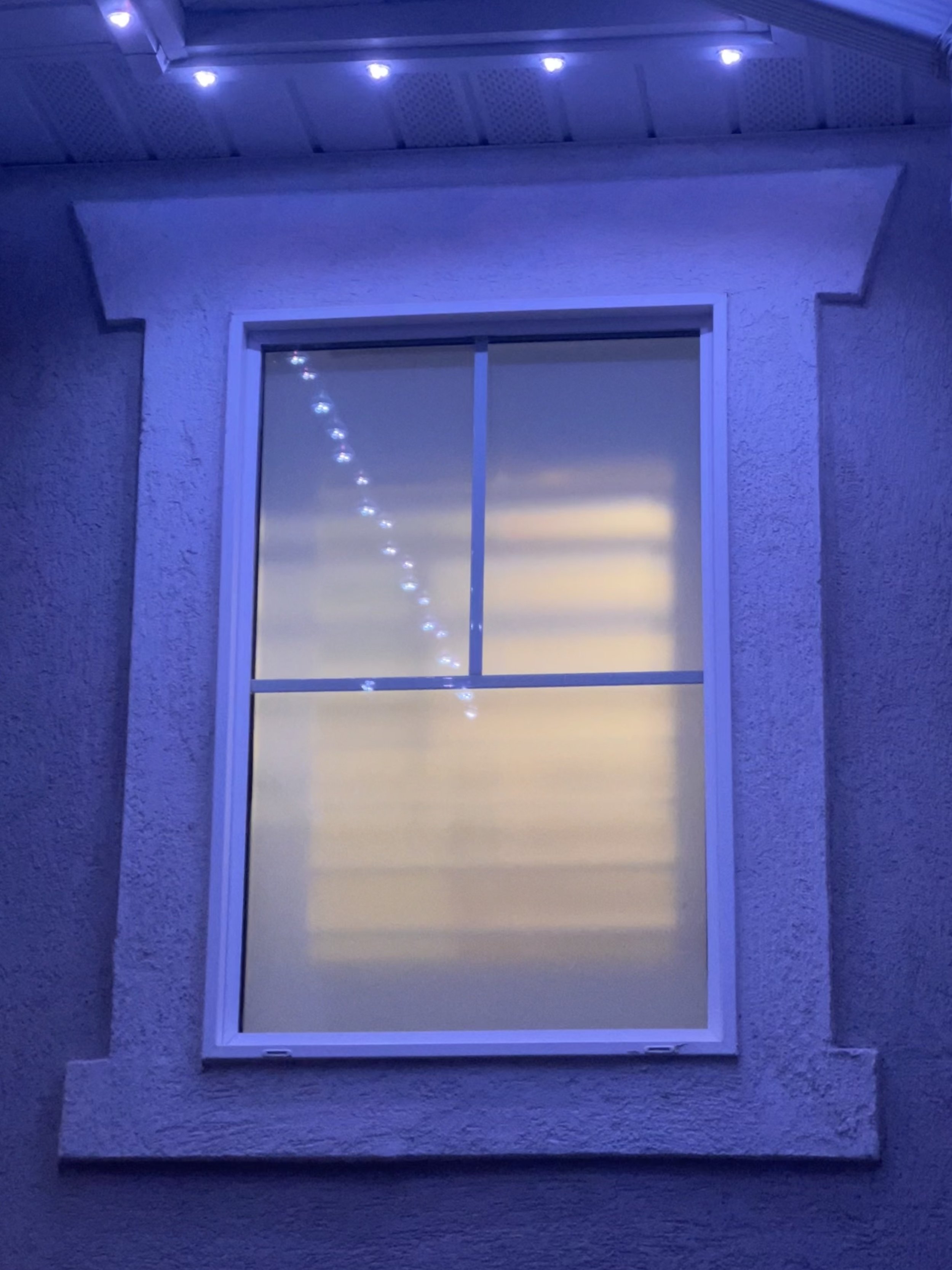 Close-up photo of a window on a building wall, lit with blue-purple exterior lighting, with a string of small lights reflected on the glass.