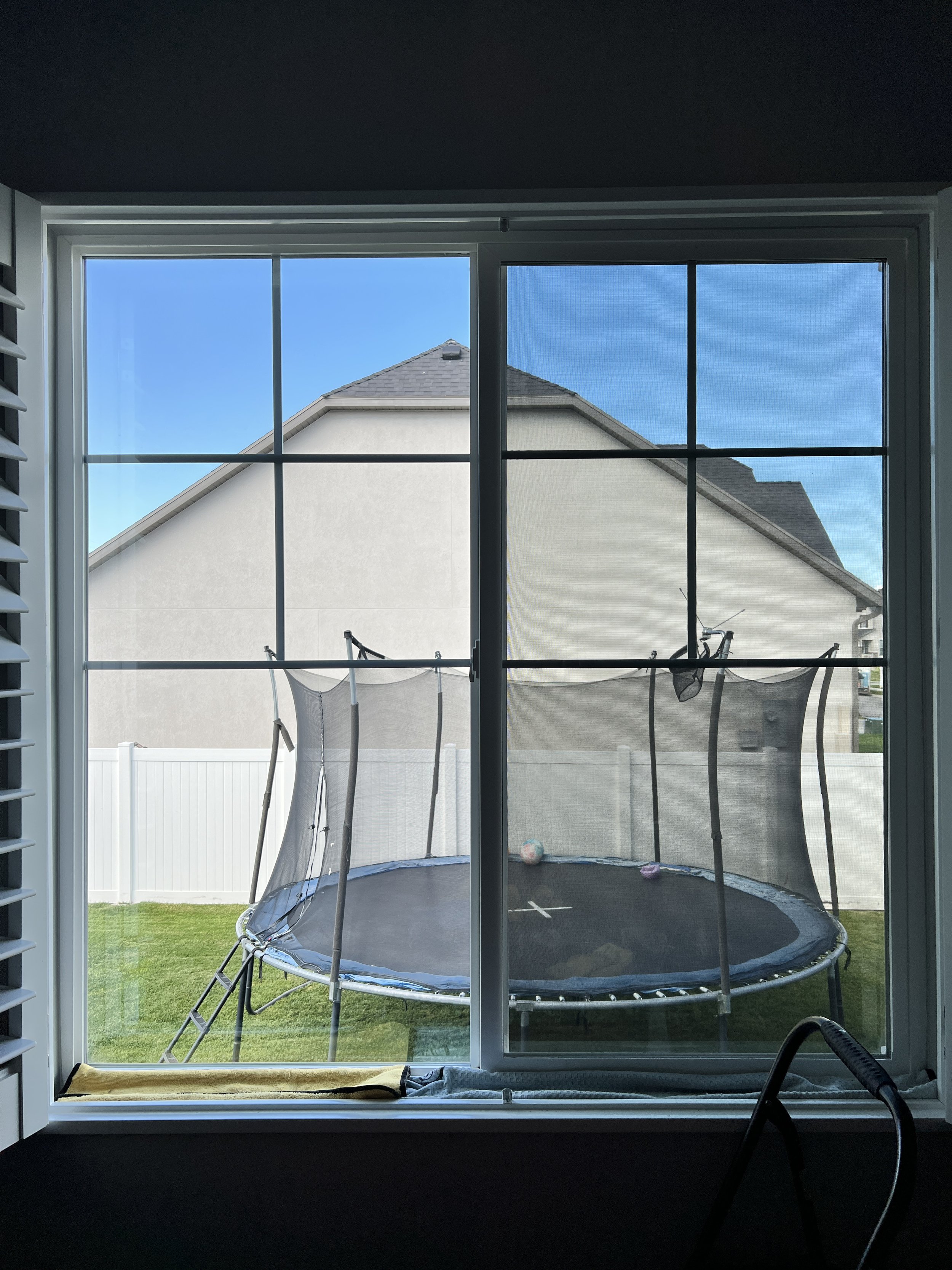 View through a window showing a backyard with a trampoline and a white fence, neighboring houses, and a blue sky.