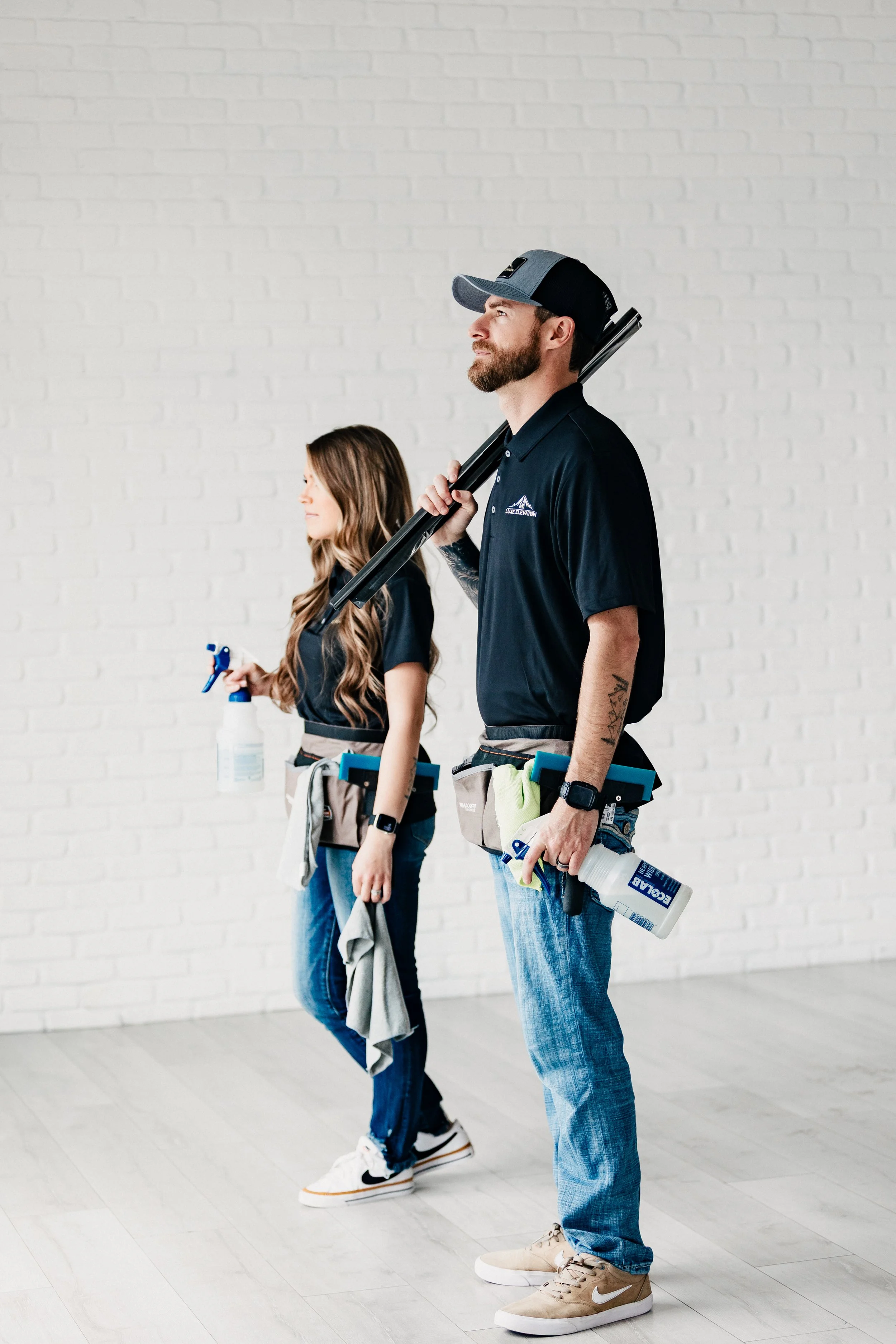 Two individuals, a man and a woman, standing side by side, holding cleaning tools, with the man carrying a long tool over his shoulder and the woman holding a spray bottle. Both are wearing dark polo shirts and pants, with tool belts around their waists, against a white brick wall background.