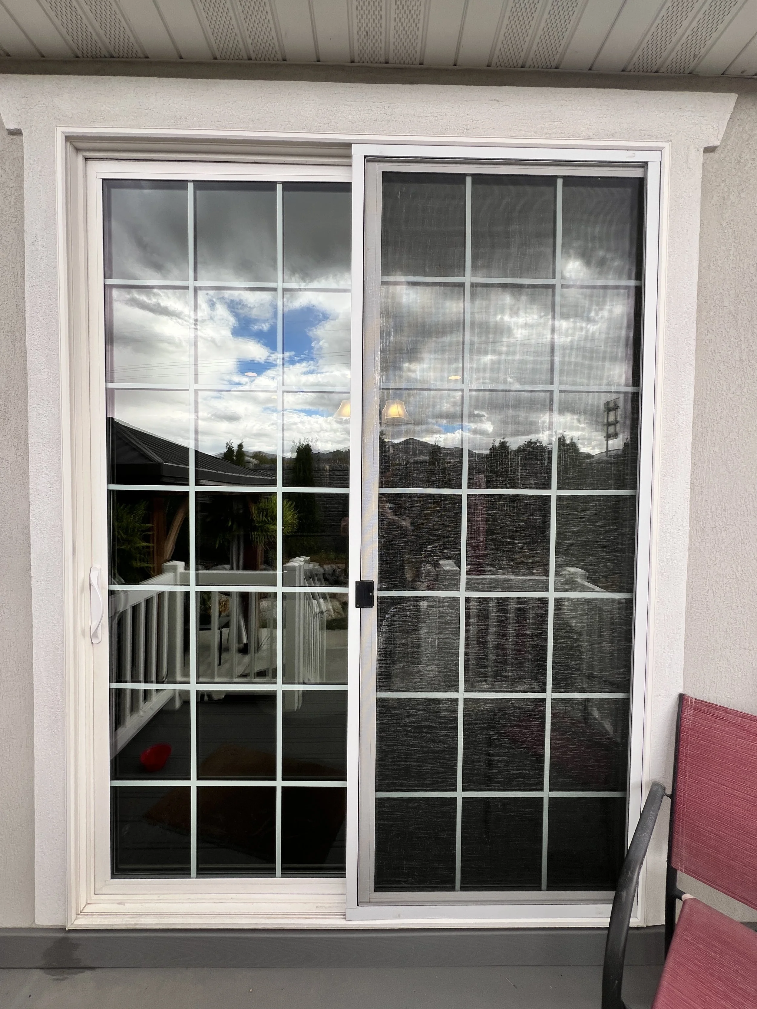 Sliding glass door with grid pattern, reflecting a cloudy sky and outdoor scenery, with a red chair on the right.