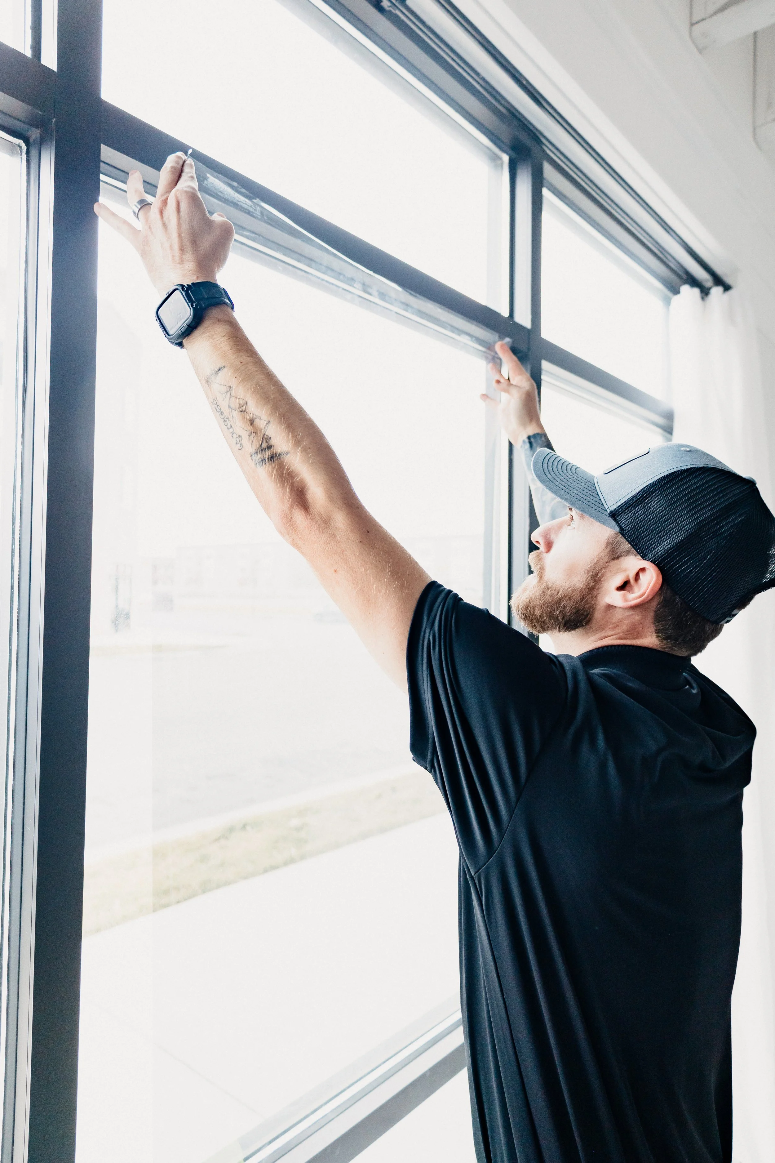 A man wearing a black cap, black shirt, and smartwatch is installing or adjusting a window screen inside a building with white curtains and large windows.