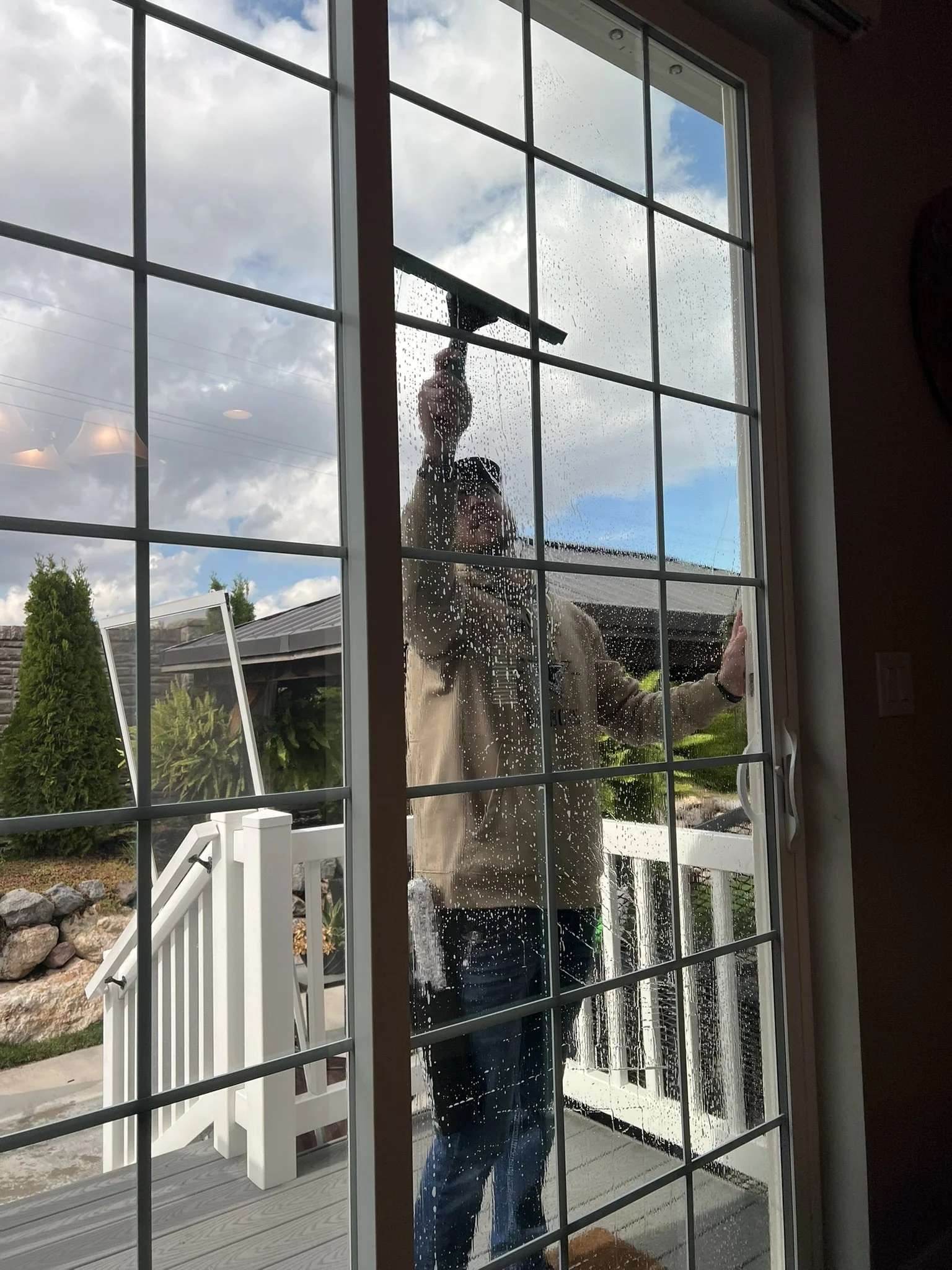Person cleaning a glass door from outside, wiping the glass with a squeegee while the sun is shining and rain droplets are visible on the glass.