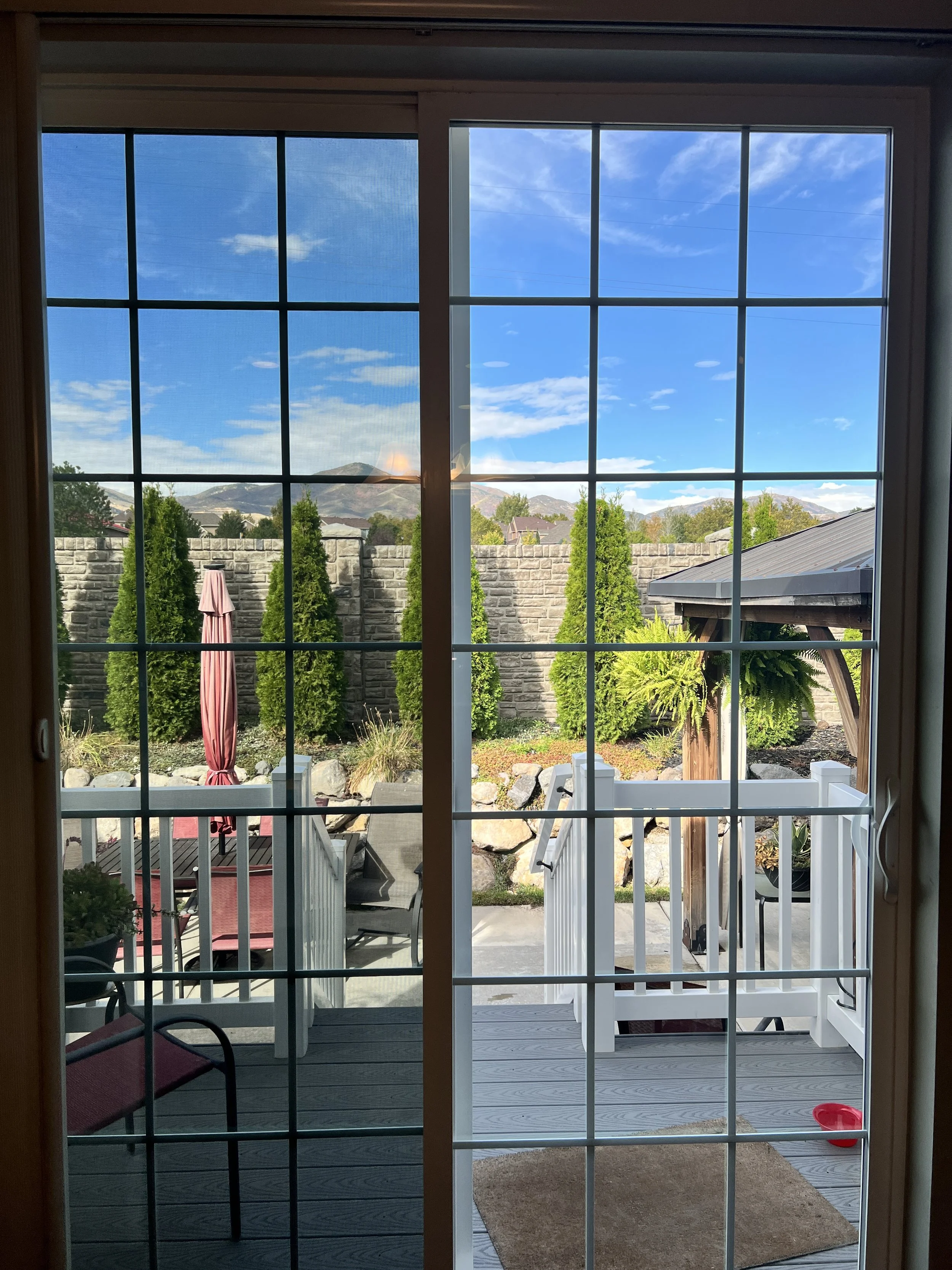 View through a sliding glass door onto a backyard patio with outdoor furniture, a closed pink umbrella, green trees, a stone wall, and mountains in the background under a partly cloudy sky.