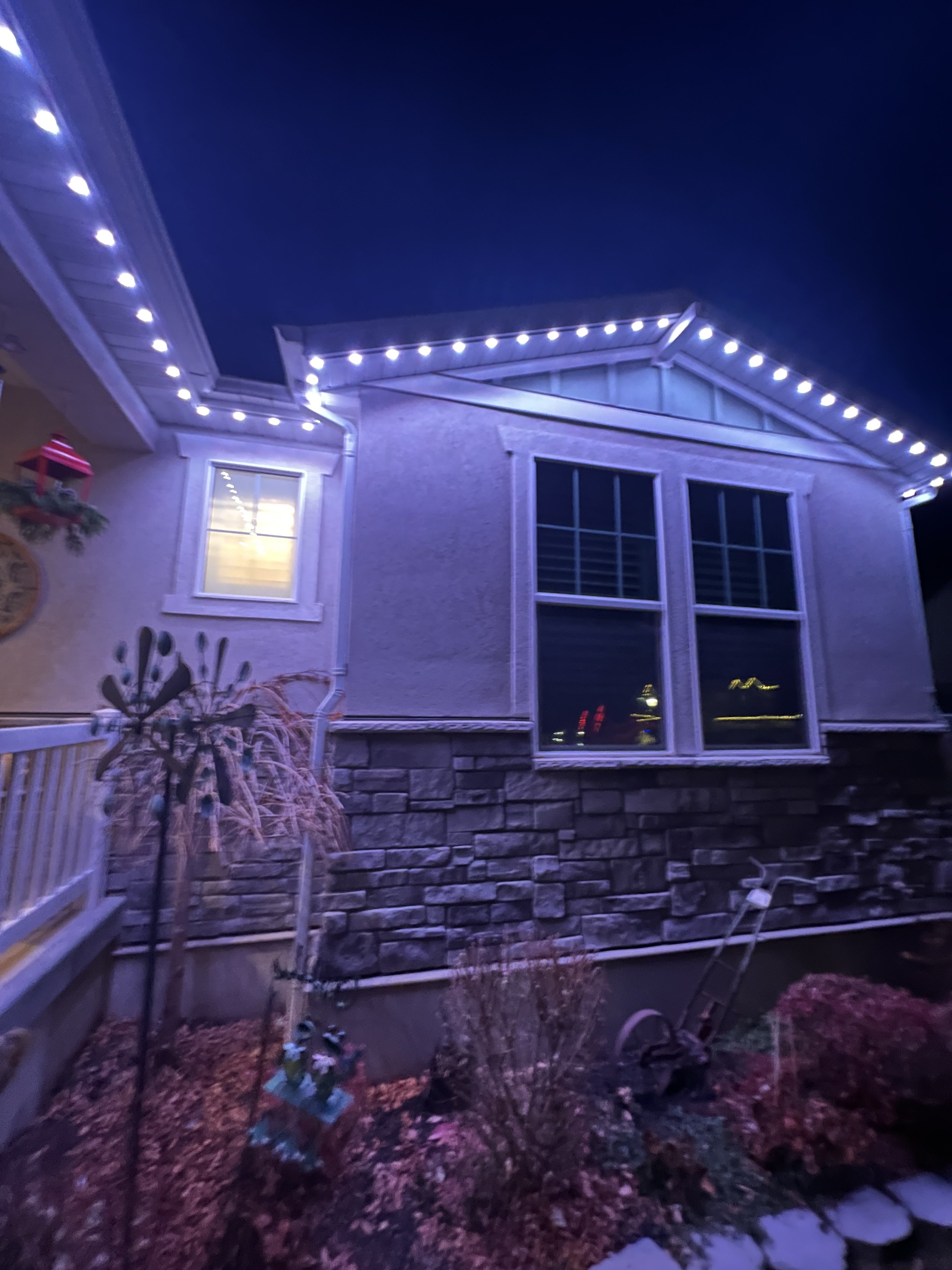 House exterior at night decorated with white string lights along the roofline and a lit window. The garden includes stone and wood elements, with bushes, a small tree, and decorative items.