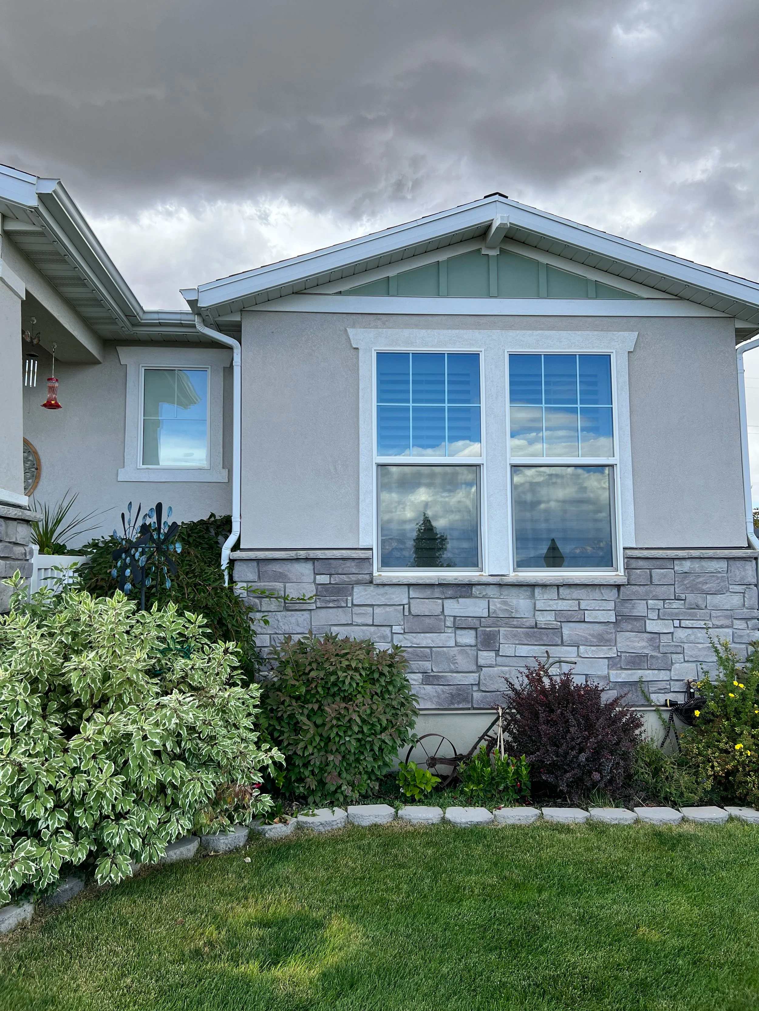 Front view of a house with a stone facade, large double-hung window, and a well-maintained lawn with shrubs and plants under a cloudy sky.