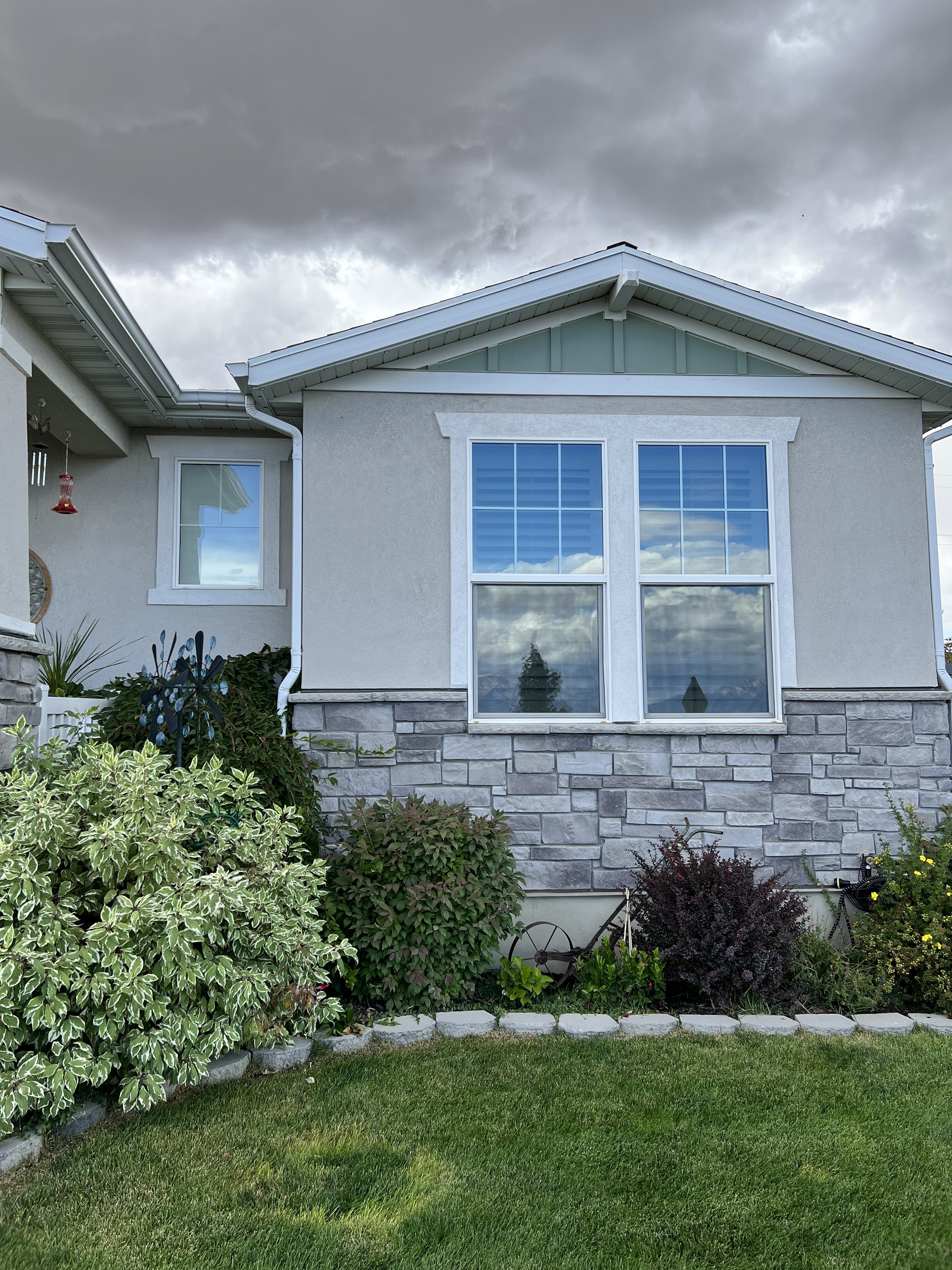 Photo of a house with gray stone foundation, white exterior walls, and large windows reflecting clouds and sky. The front yard has green grass and assorted bushes and plants. Dark clouds are present in the sky.