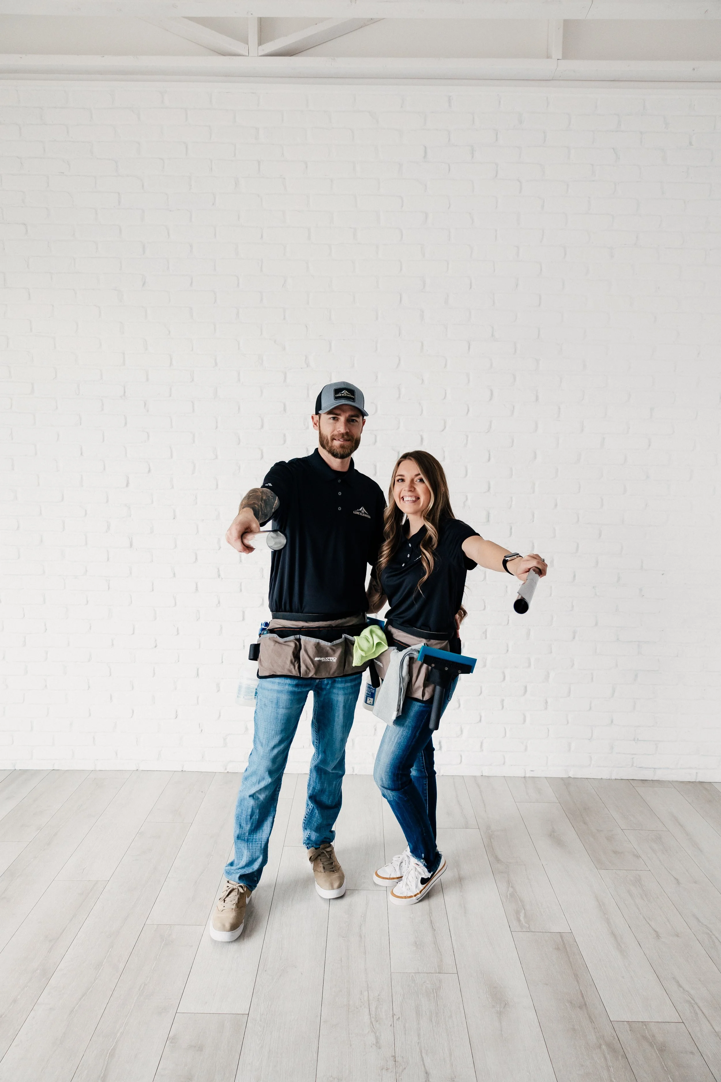 Two smiling people wearing work belts and holding cleaning tools stand in front of a white brick wall, posing with arms extended outward.