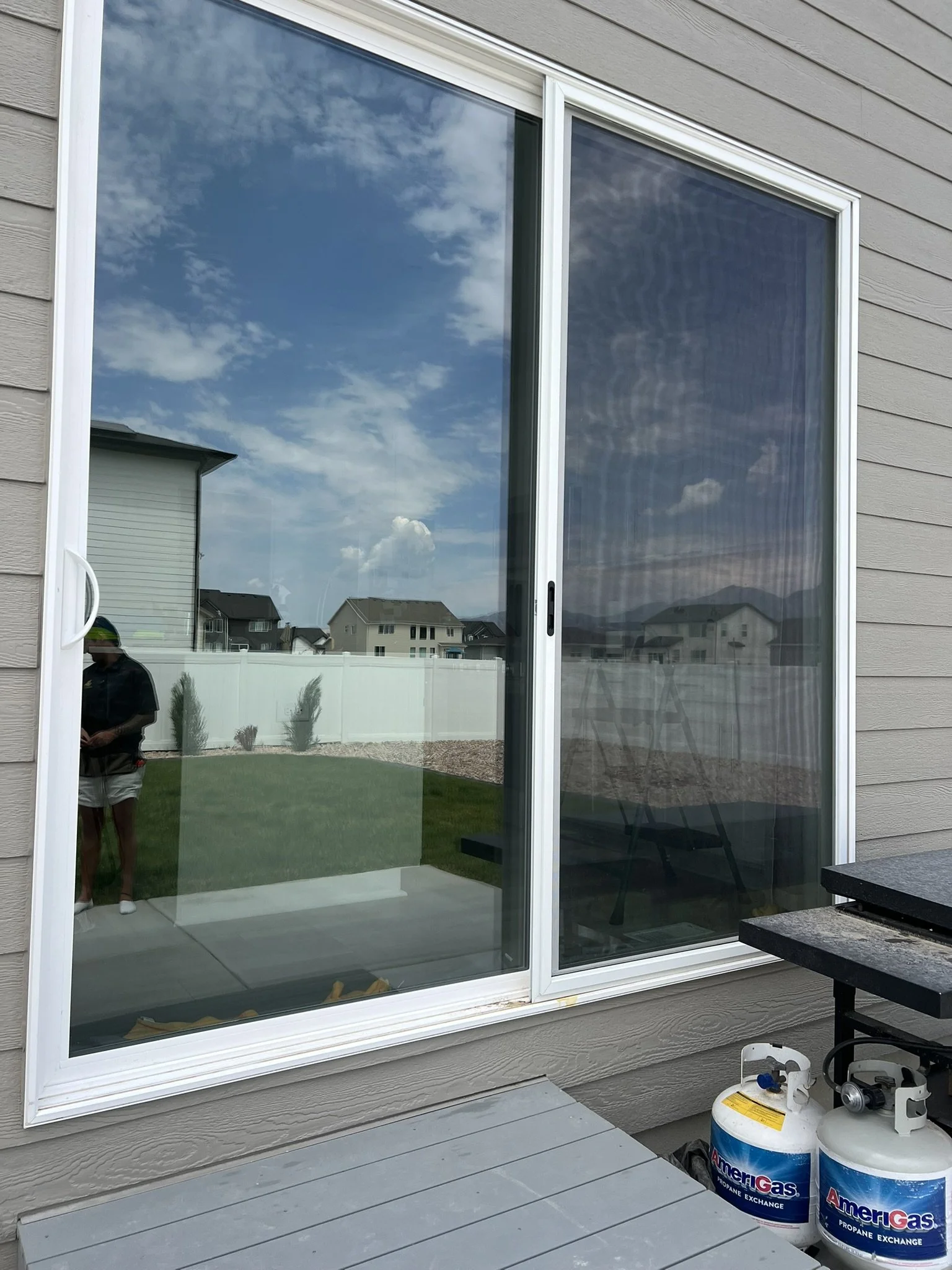 View of a sliding glass door with a reflection of the sky, clouds, and neighboring houses, and a person standing outside.