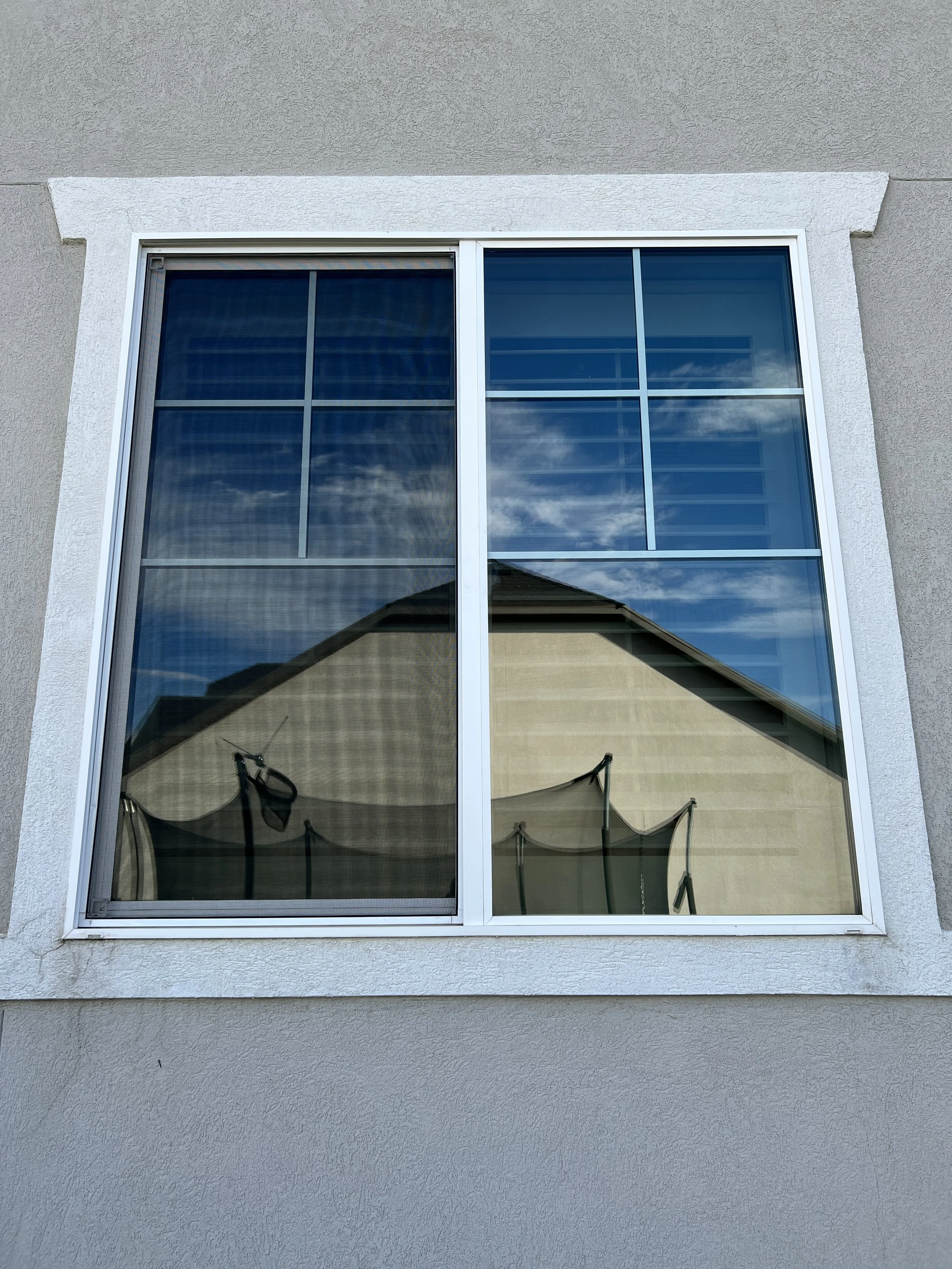 A house window with reflections of the sky, clouds, a roof, patio furniture, and an outdoor umbrella.