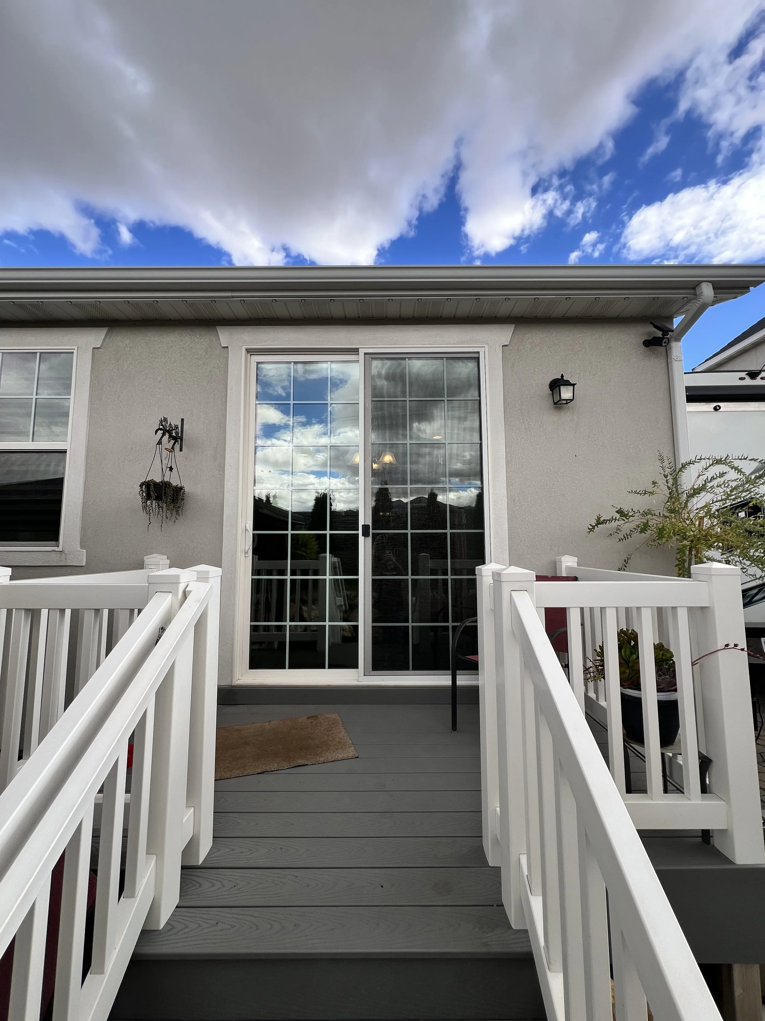 View of a house patio with white railing, gray wooden deck, glass sliding door, and exterior wall lights, under a partly cloudy sky.