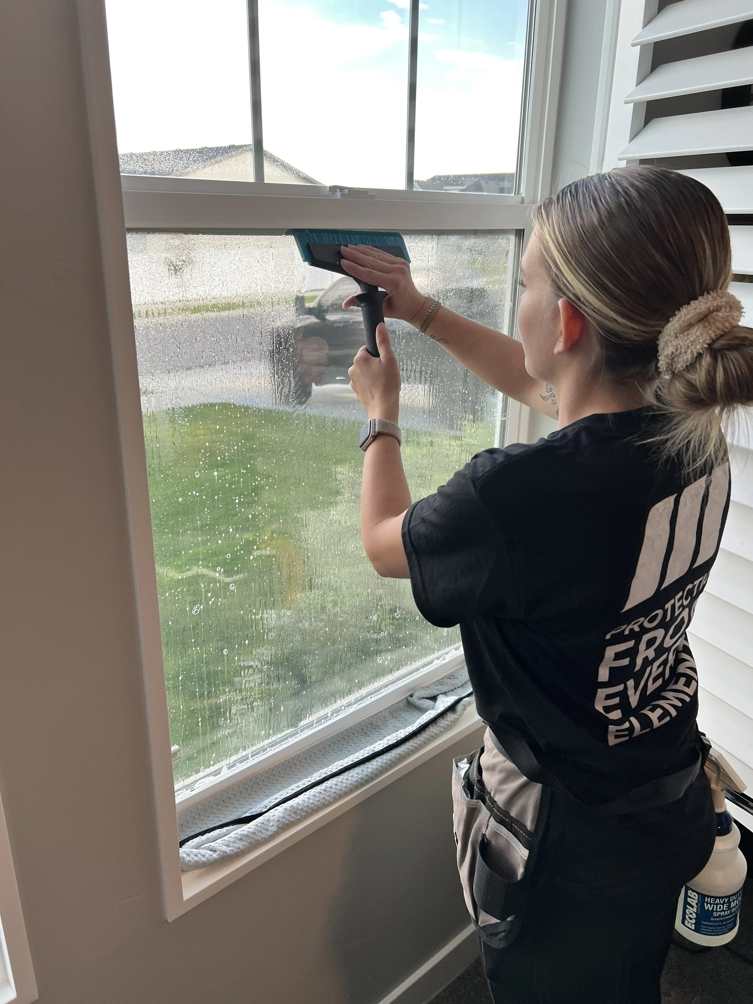 A woman cleaning a large window with a squeegee, water droplets on the glass, outside view of a car, grass, and cloudy sky.