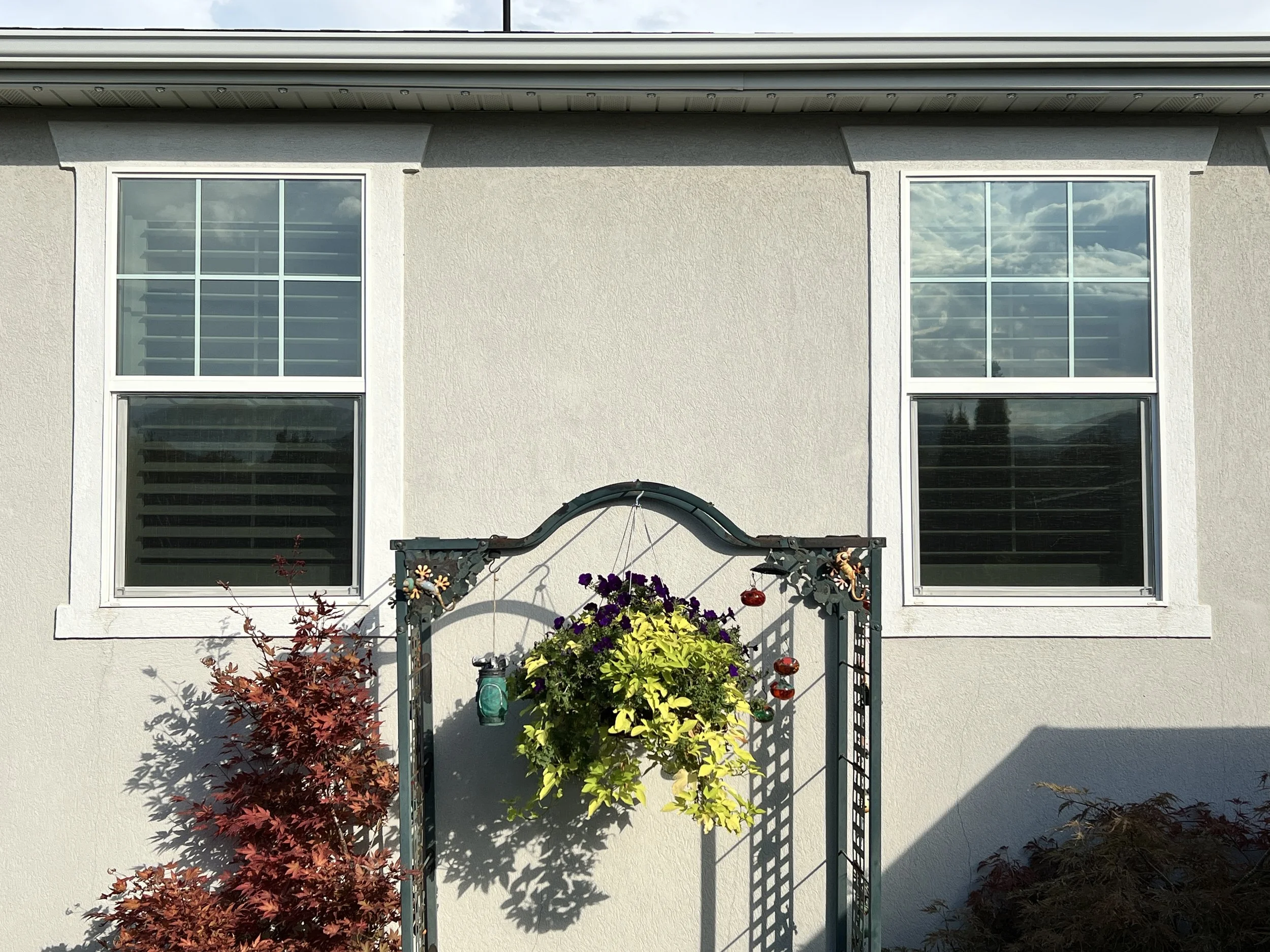 A beige house with two large windows and a green garden archway in front, decorated with hanging ornaments and a large hanging flower pot, with bushes and trees around.