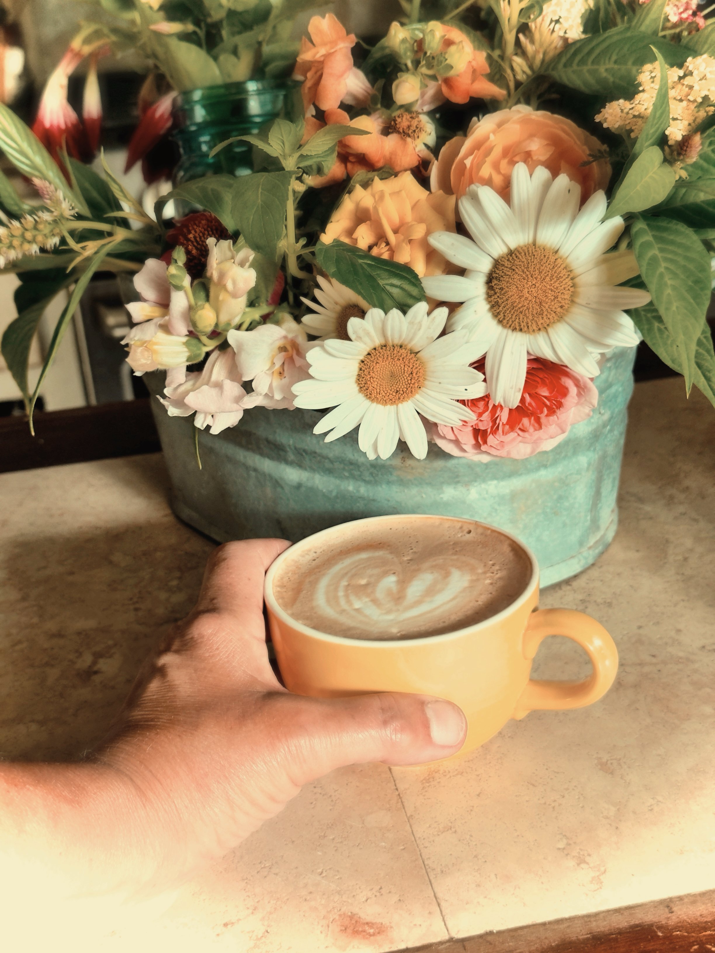 My hand holding a creamy cup of coffee in a vintage yellow mug with a heart for latté art in front of a metal tub of flowers grown here at Subherbs & blooms. Snapdragons and daisies with celosia and wildflowers.