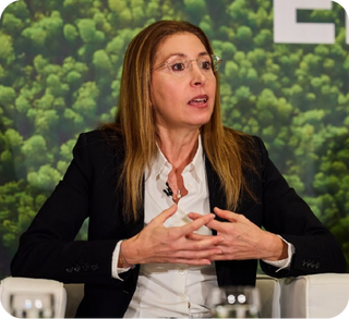 Woman with long brown hair and glasses in a black blazer and white shirt speaking at a panel.