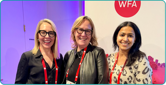 Three women smiling at an event with a red WFA logo in the background.