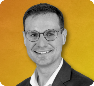Professional headshot of a man wearing glasses and a dark suit with a white shirt smiling against a yellow background.