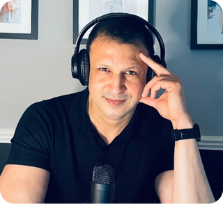 Man wearing headphones posing in front of a microphone indoors with framed pictures on the wall behind him.