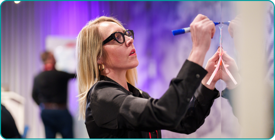 Woman with blonde hair and glasses in a black blazer writing on a wall at a workshop.