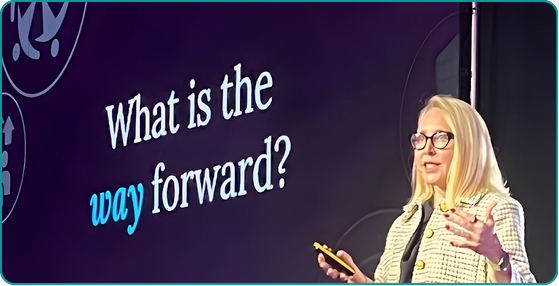 Speaker with blonde hair and glasses wearing a yellow jacket next to a large screen asking “What is the way forward?”