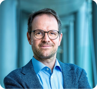Portrait of a man with glasses wearing a blue blazer and shirt in front of a blurred modern building background.