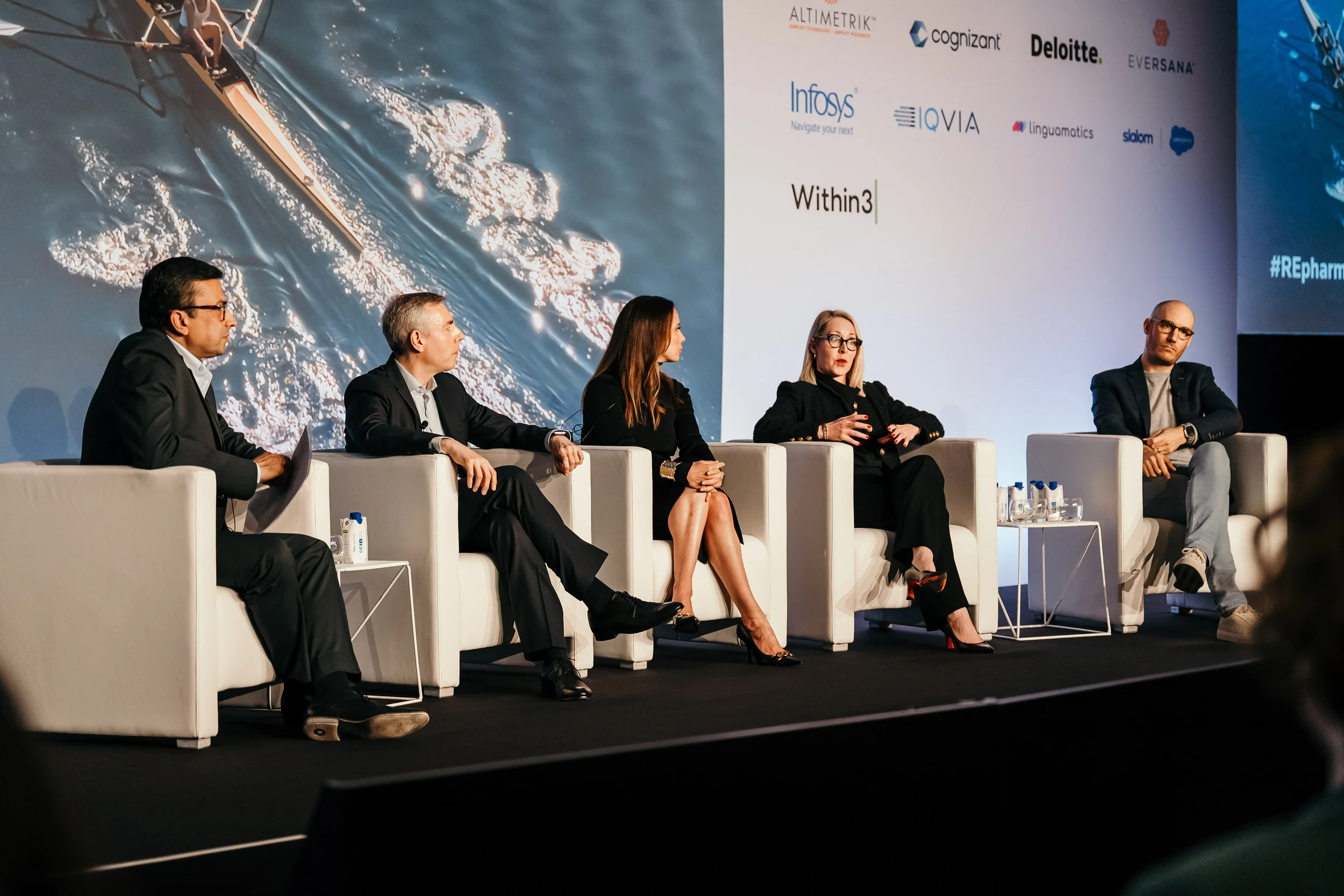 Panel of five individuals on white chairs on stage at a conference with a large screen showing logos and a boat on water.