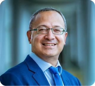A smiling man in a blue suit and glasses standing indoors with a blurred background.