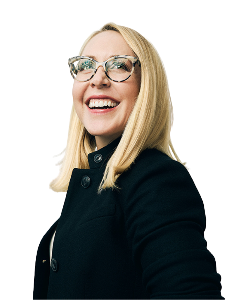 A woman with blonde hair, glasses, and a black coat smiling brightly, looking slightly upward.