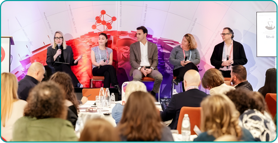 Panel of five people on stage at a conference with a cityscape backdrop and audience seated at tables.
