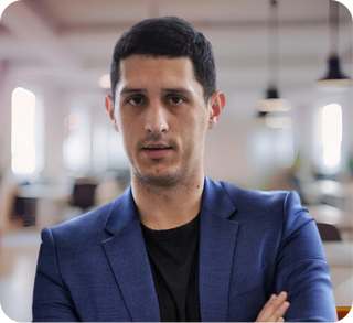 Man with short dark hair wearing a blue blazer and black shirt standing in a bright indoor space with a blurred background.