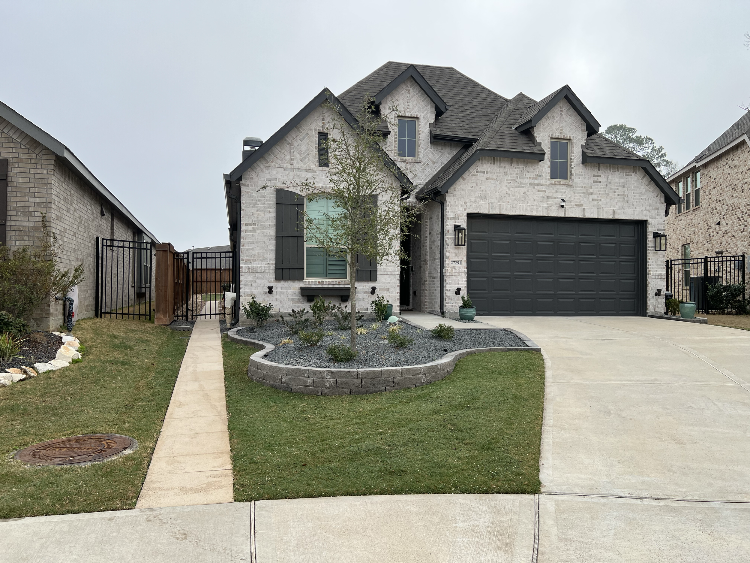 A picture of a Slim brand new white brick suburban home with black accents with a gray rock landscape and a perfect small green lawn with no weeds contrasting to a neighboring home uneven with weeds. 