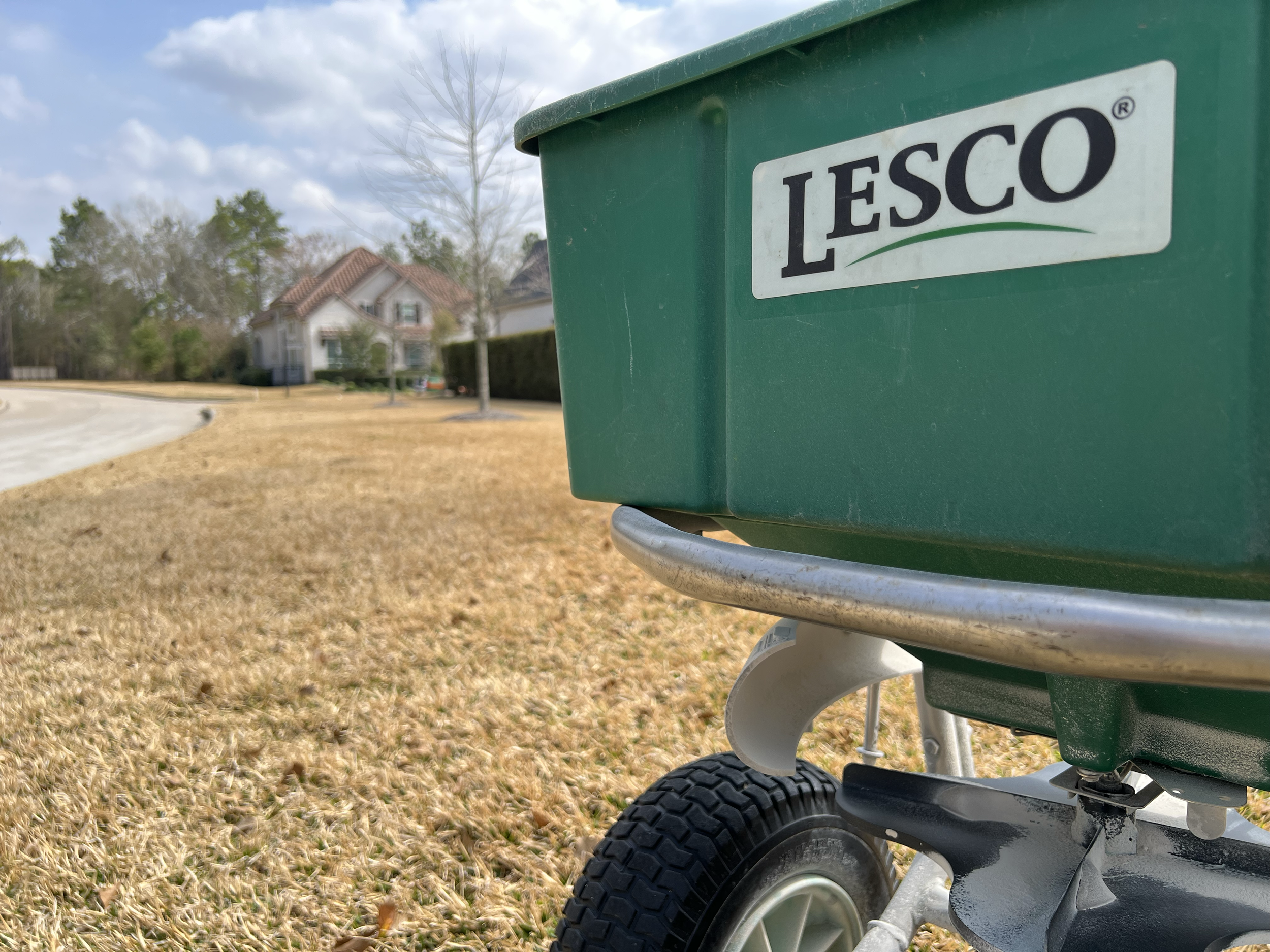 A picture of a Lesco spreader close up shot of the front of the hopper logo with a dormant light brown Bermuda lawn in the background frost bitten from winter.