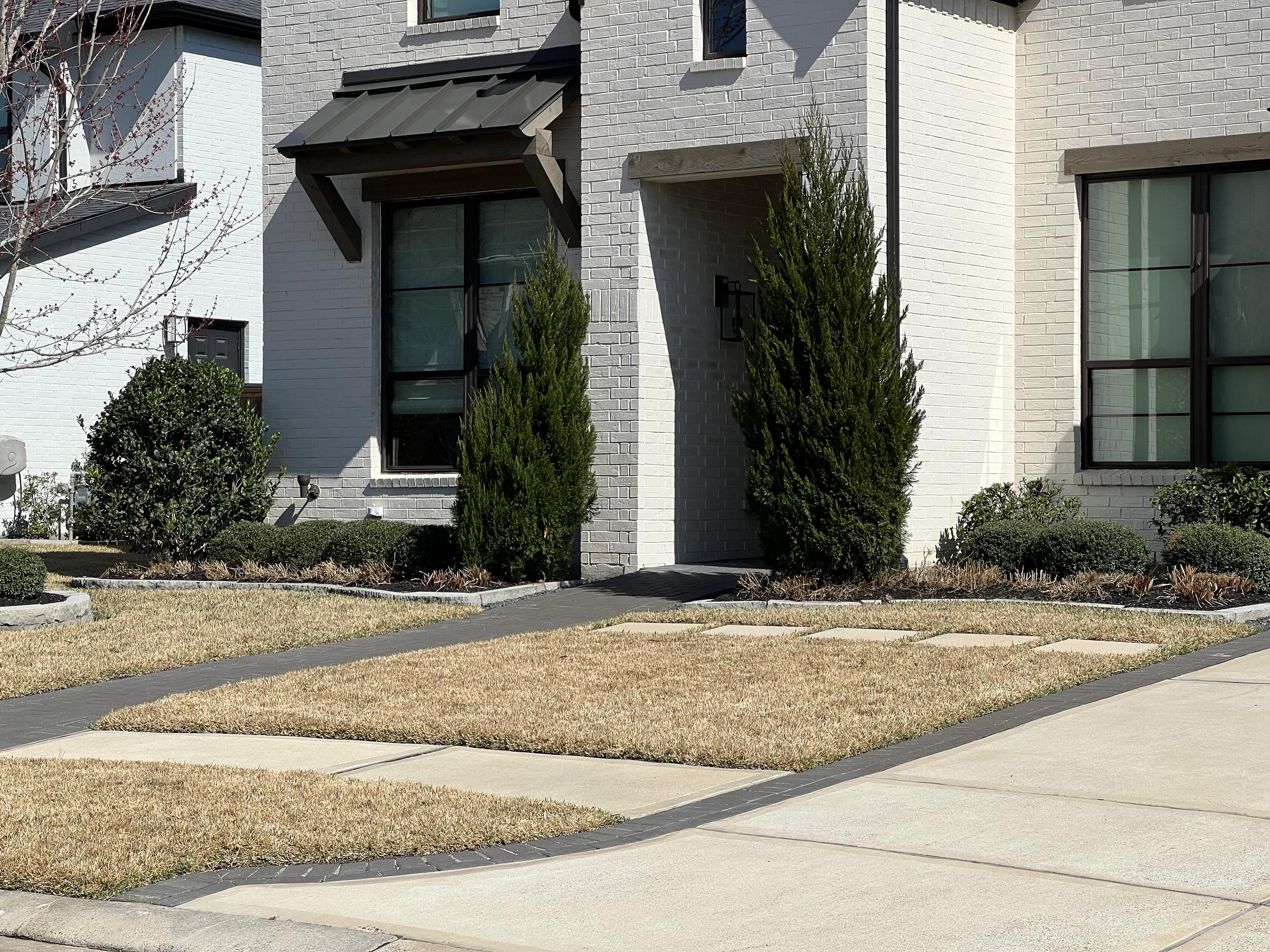 The front of a clean 2 story white suburban home in the outskirts of Houston tx with black framed windows sporting an evergreen landscape and perfectly even colored dormant Bermuda lawn with no weeds or uneven coloring. 