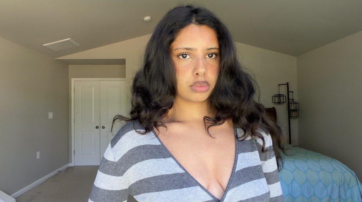 A woman with long wavy black hair wearing a gray and white striped top standing in a bedroom with beige walls and closed double doors.