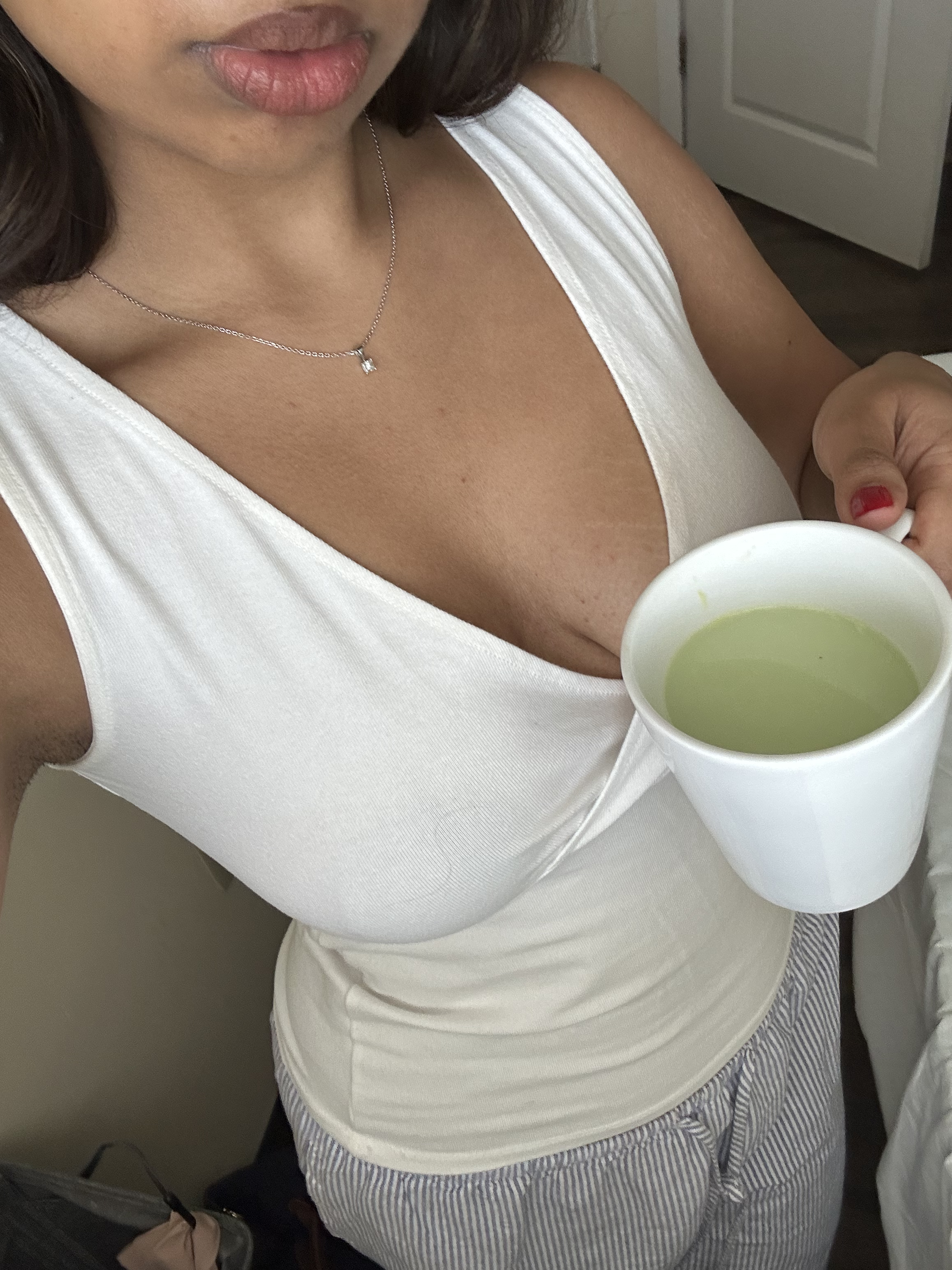 A woman wearing a white sleeveless top, a delicate necklace, and striped pants, holding a white mug filled with a yellow-green beverage.