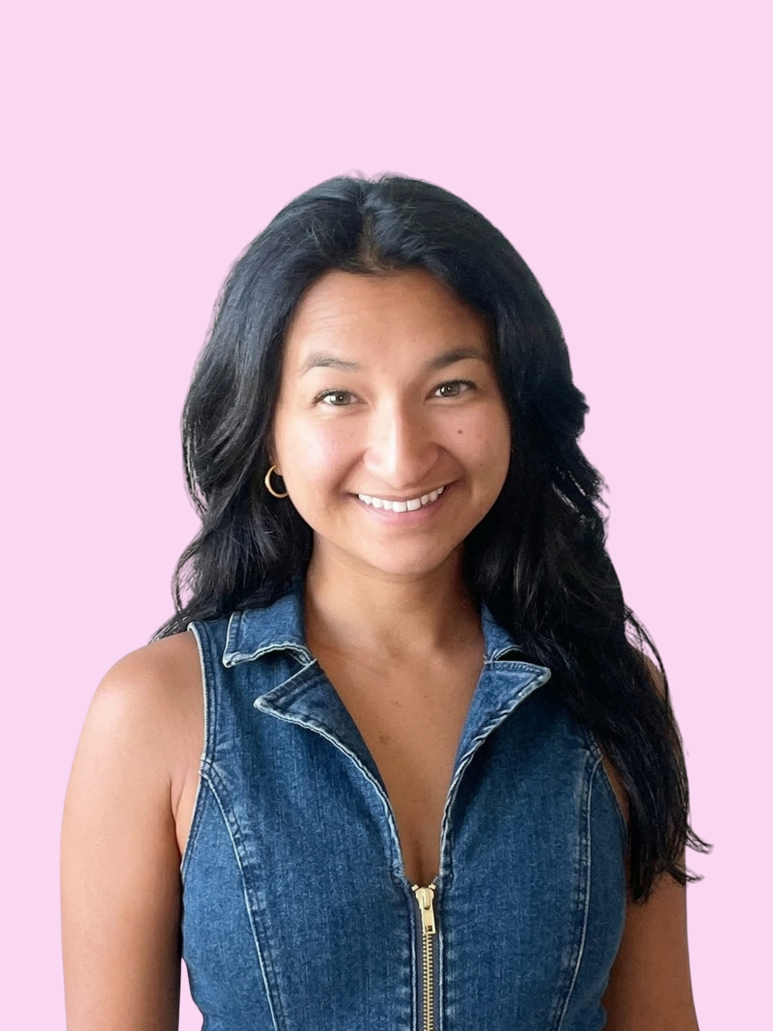 A woman with long dark hair smiling, wearing a sleeveless denim jacket against a light pink background.