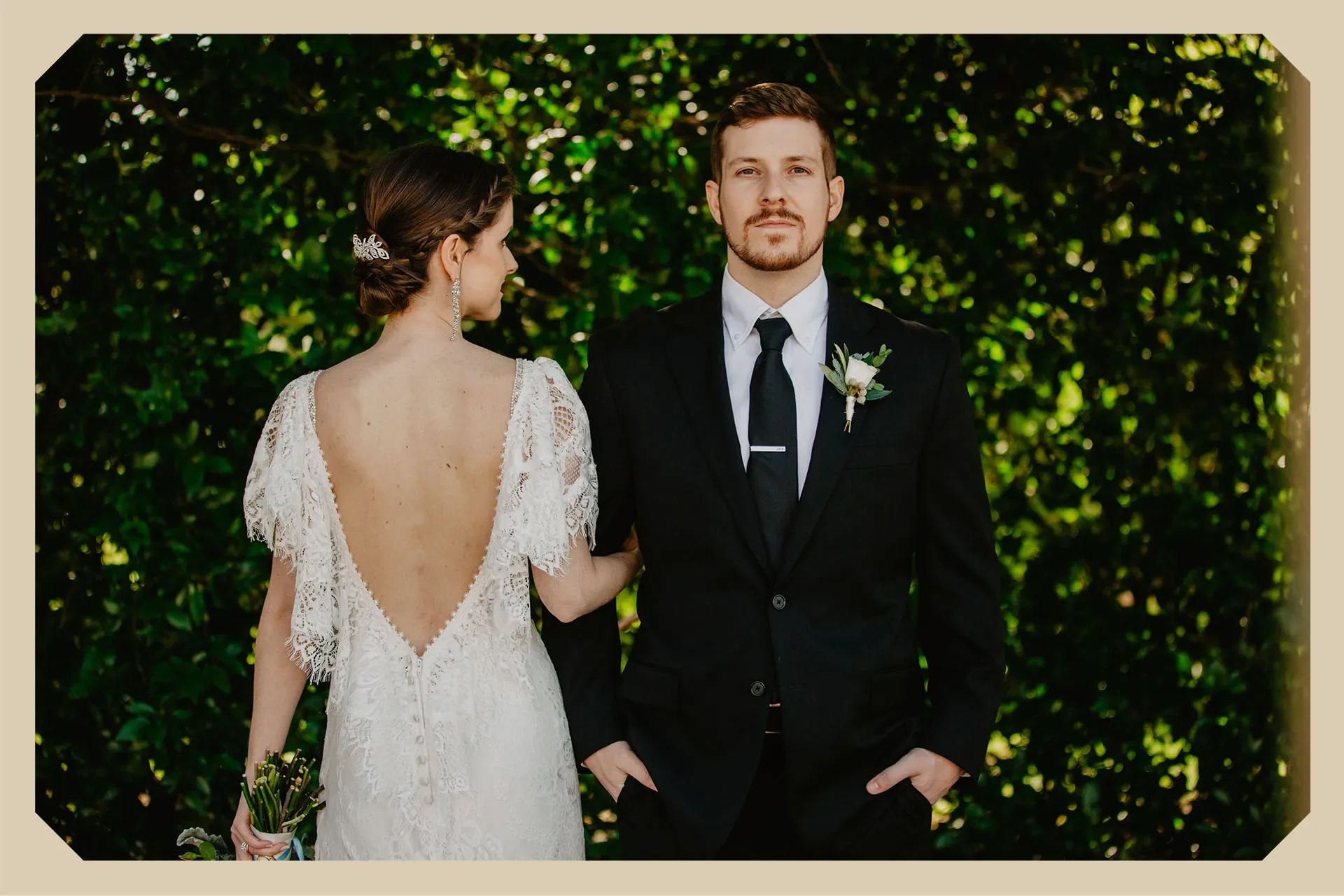 A couple posed dramatically, outdoors in front of green foliage. The bride is turned to the side with an open back wedding dress and holding a small bouquet. The groom is facing forward, dressed in a black suit & with a white shirt and a boutonniere.