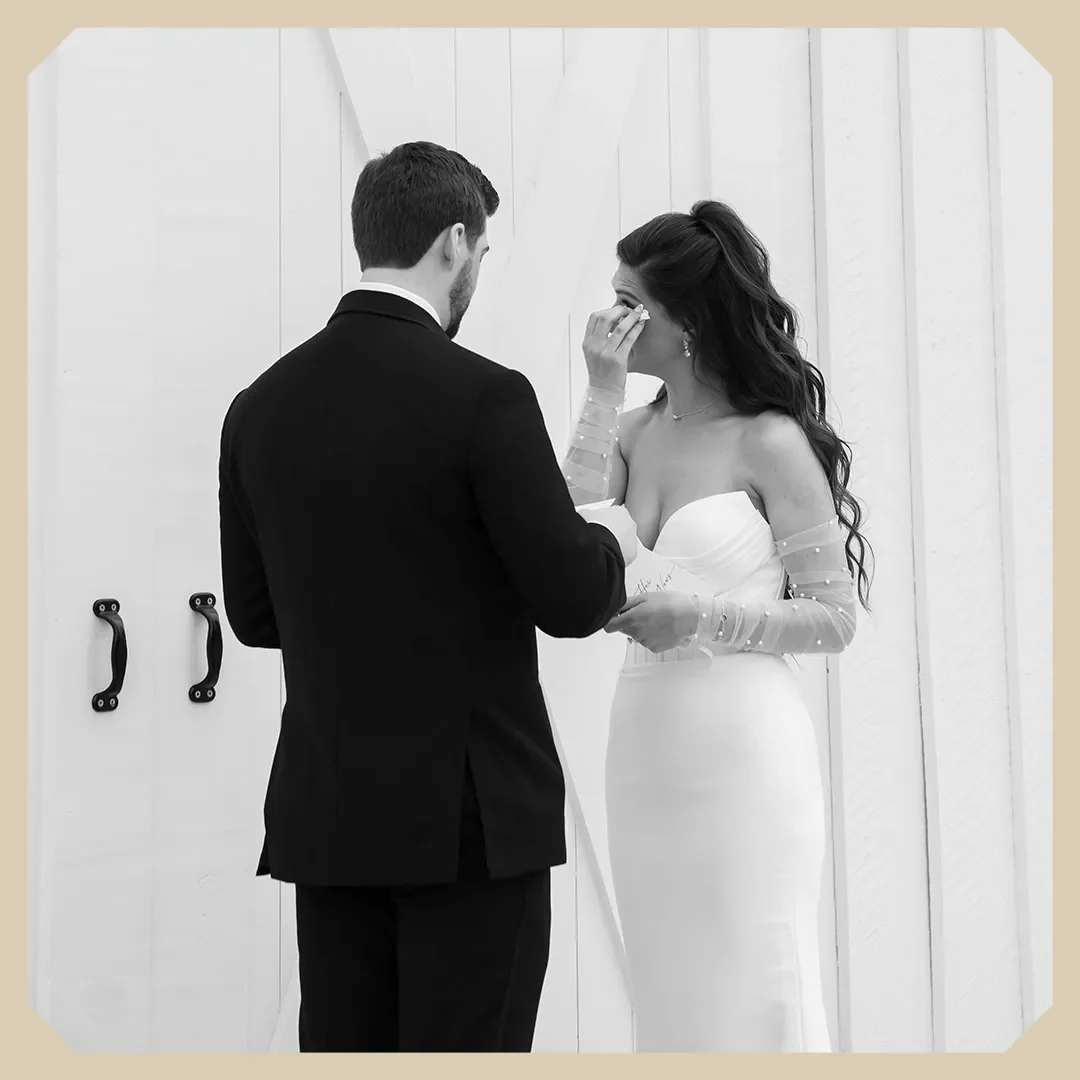 A bride crying during her wedding vows as the groom holds her hands, standing in front of a white paneled barn