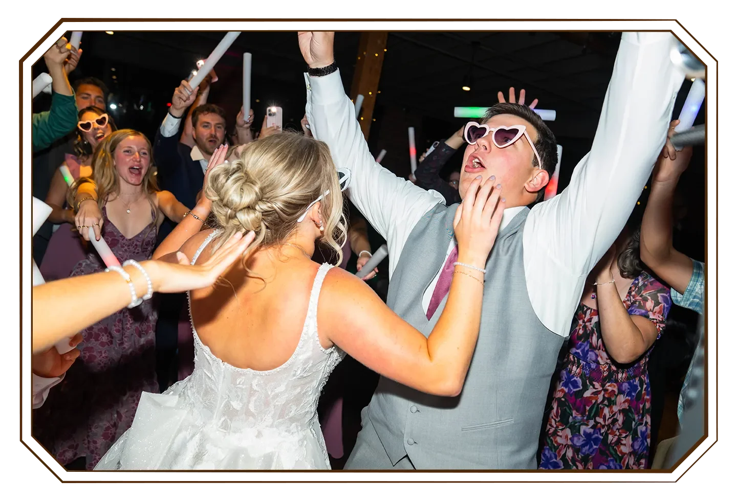 A newly wed couple laughing on the dance floor, surrounded by people dancing and celebrating at a party, wearing colorful outfits and sunglasses with glowing sticks in a lively indoor setting.