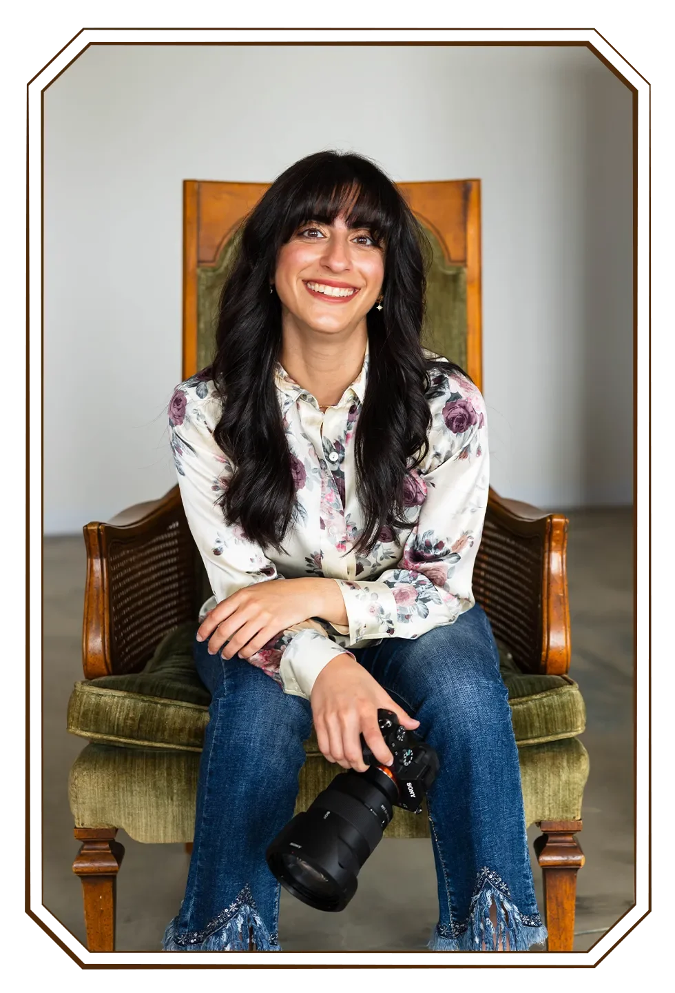 A smiling woman with long dark hair and bangs, sitting on an antique wooden and green upholstered chair, holding a professional camera in her lap, wearing a floral blouse and jeans, indoors with a clean, modern industrial background.