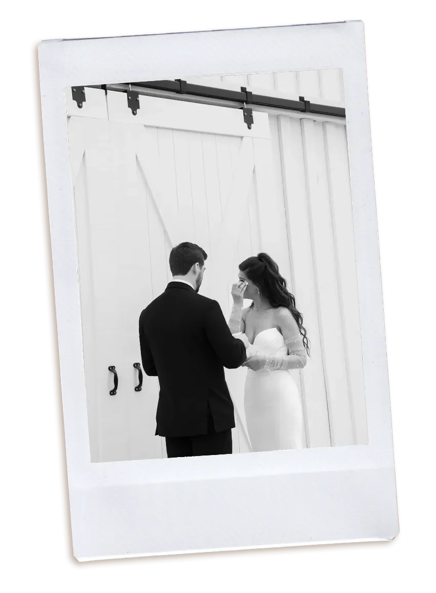 A couple dressed in wedding attire standing closely in front of sliding barn doors, with the bride drying her eyes while her groom embraces her.