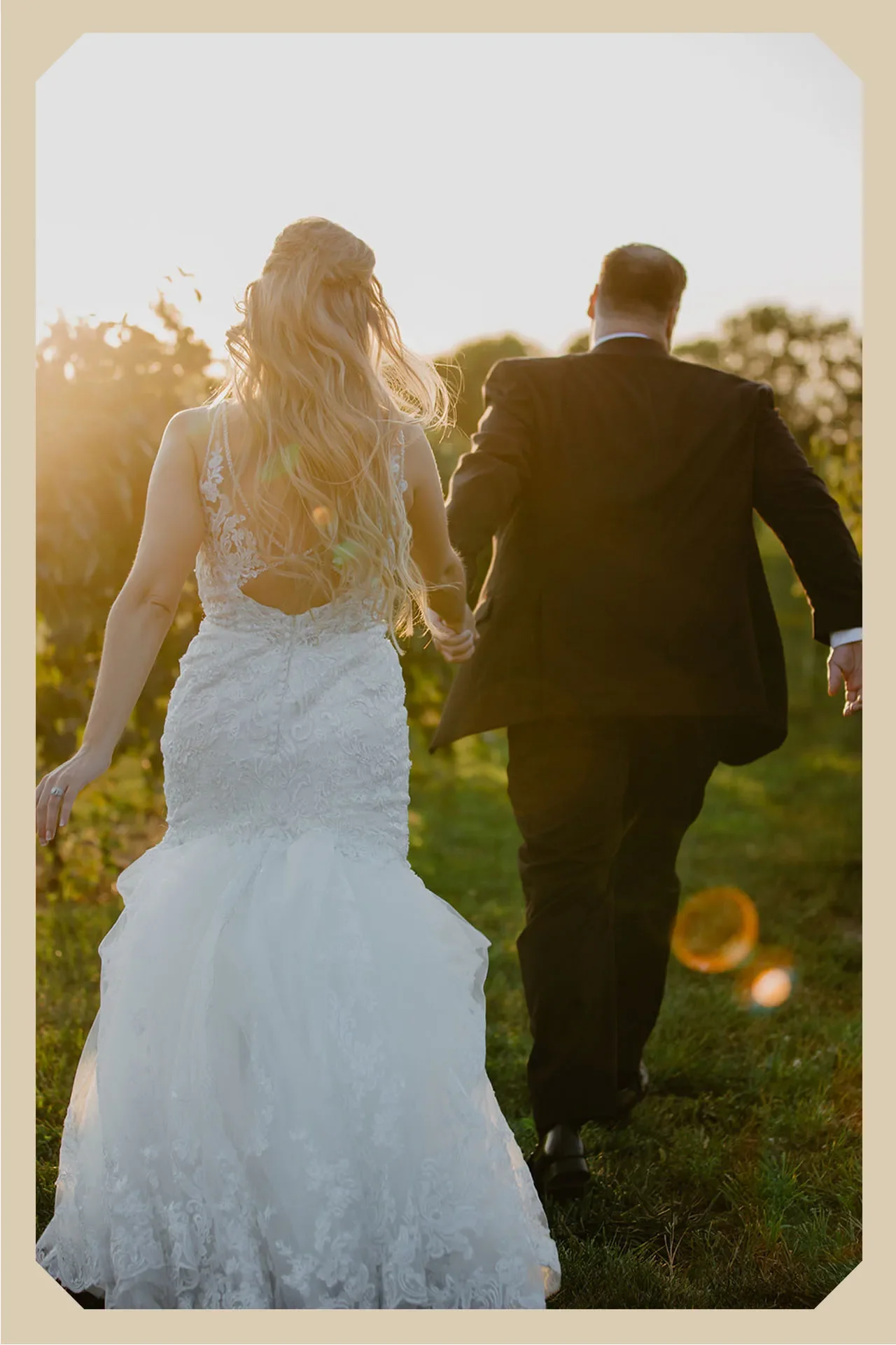 A bride and groom walking hand in hand outdoors at sunset, with the bride in a white lace wedding gown and the groom in a black suit.