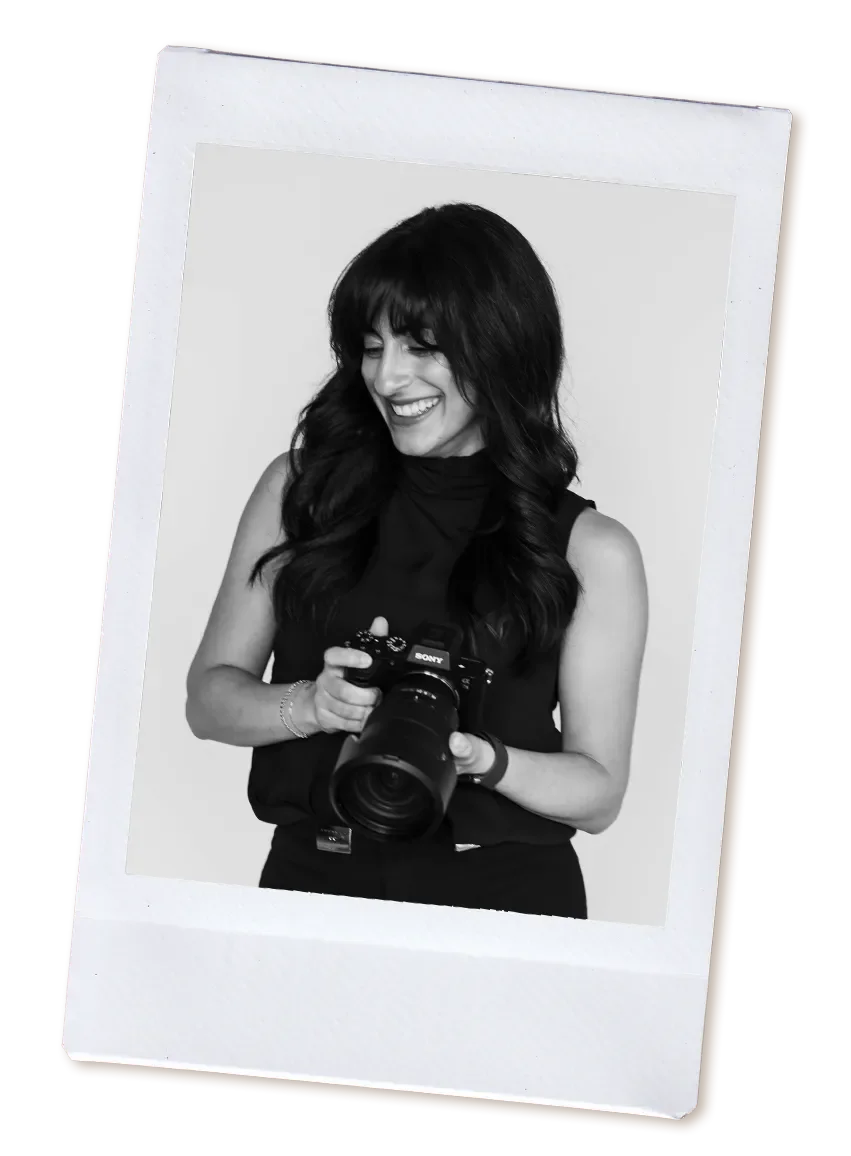 Black and white photo of a woman with dark wavy hair holding a camera, smiling and looking down.