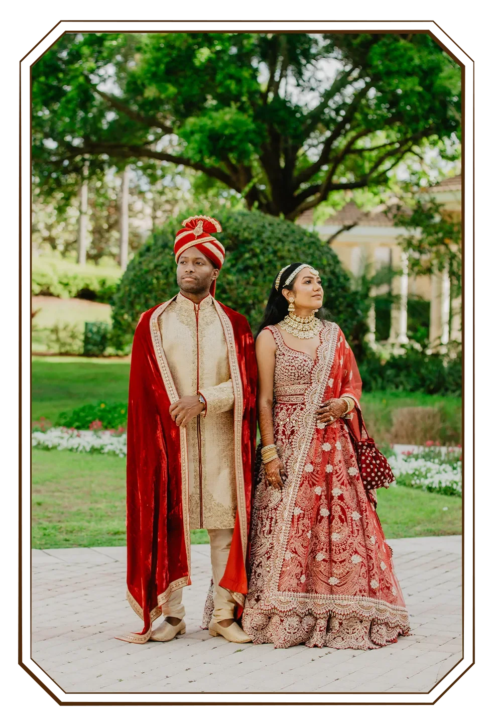 A couple dressed in traditional Indian wedding attire standing outdoors in a garden with trees and flowers.