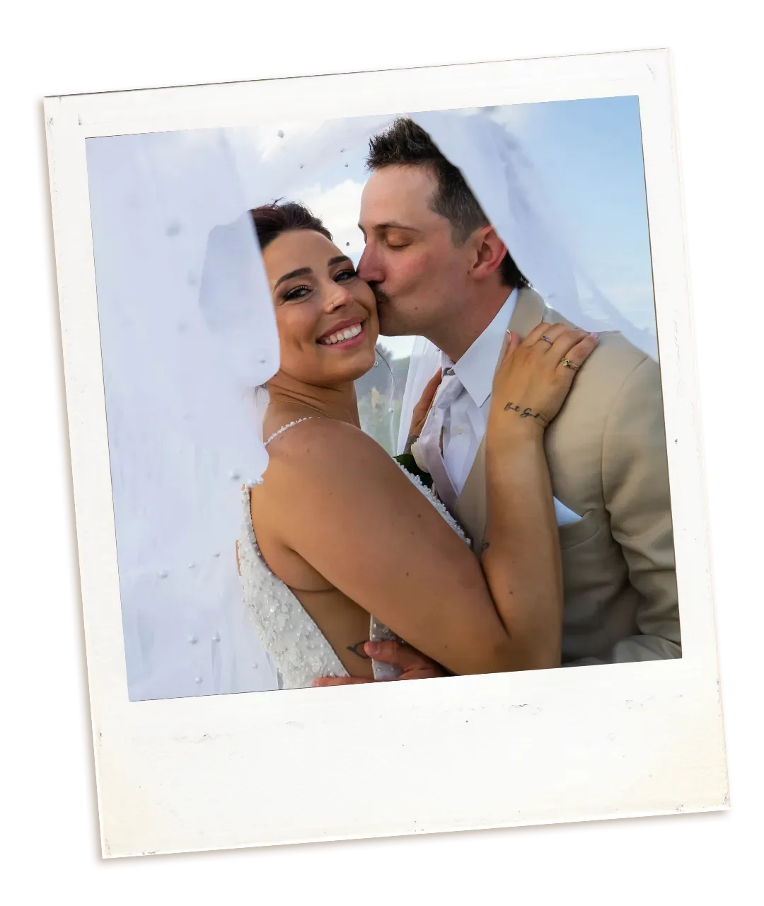 A newlywed couple embraces and kisses outside under a cloudy sky, with the veil creatively draped and framing them.