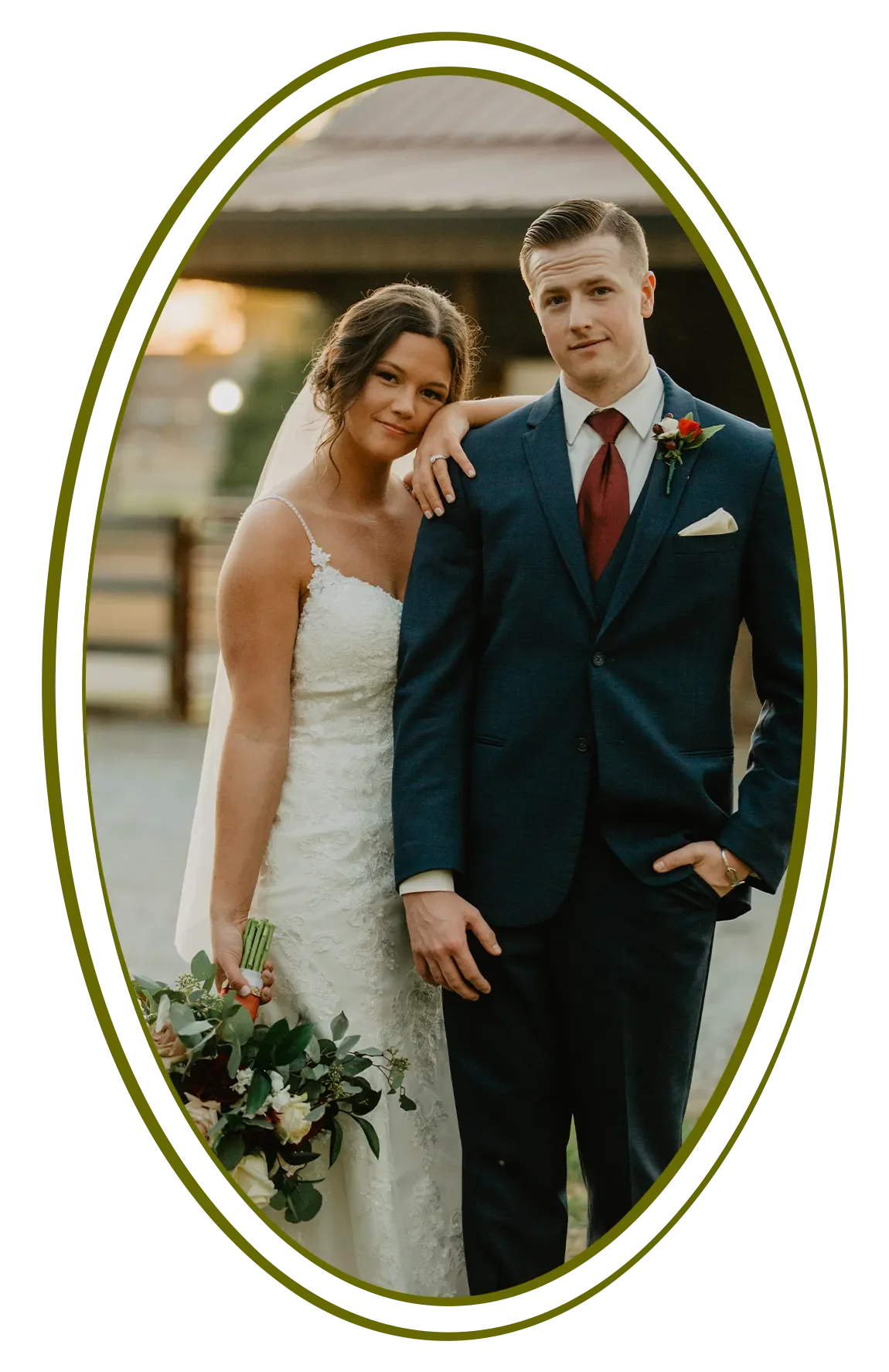 A bride and groom posing outdoors during sunset, with the bride holding a bouquet and wearing a white lace wedding dress, and the groom dressed in a navy suit with a maroon tie and boutonniere.