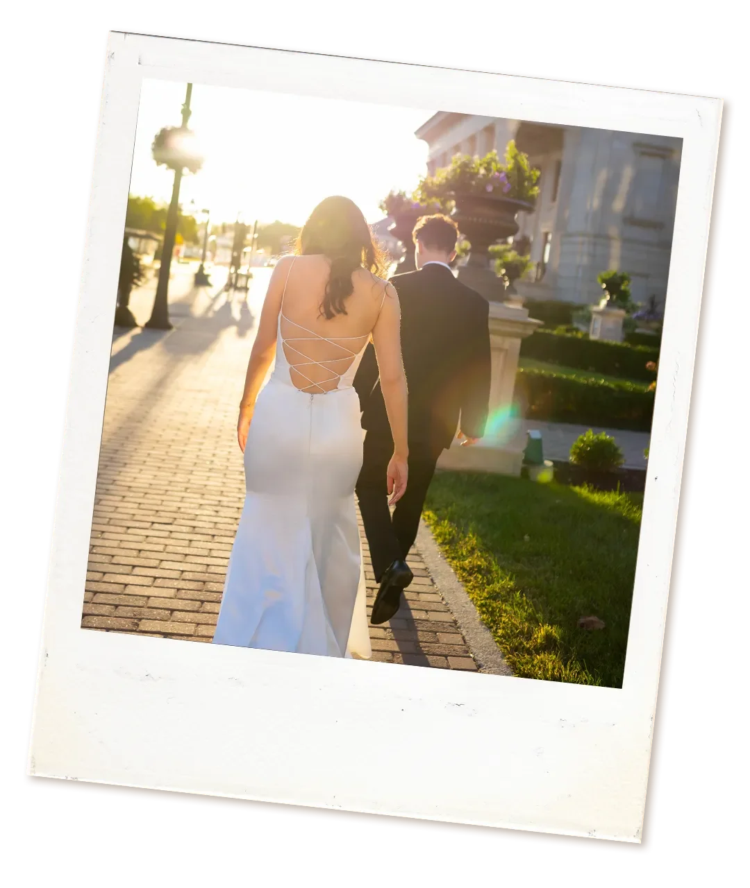A bride and groom walk hand in hand on a sidewalk during sunset, with the bride wearing a white wedding dress with an open back and the groom in a black suit.