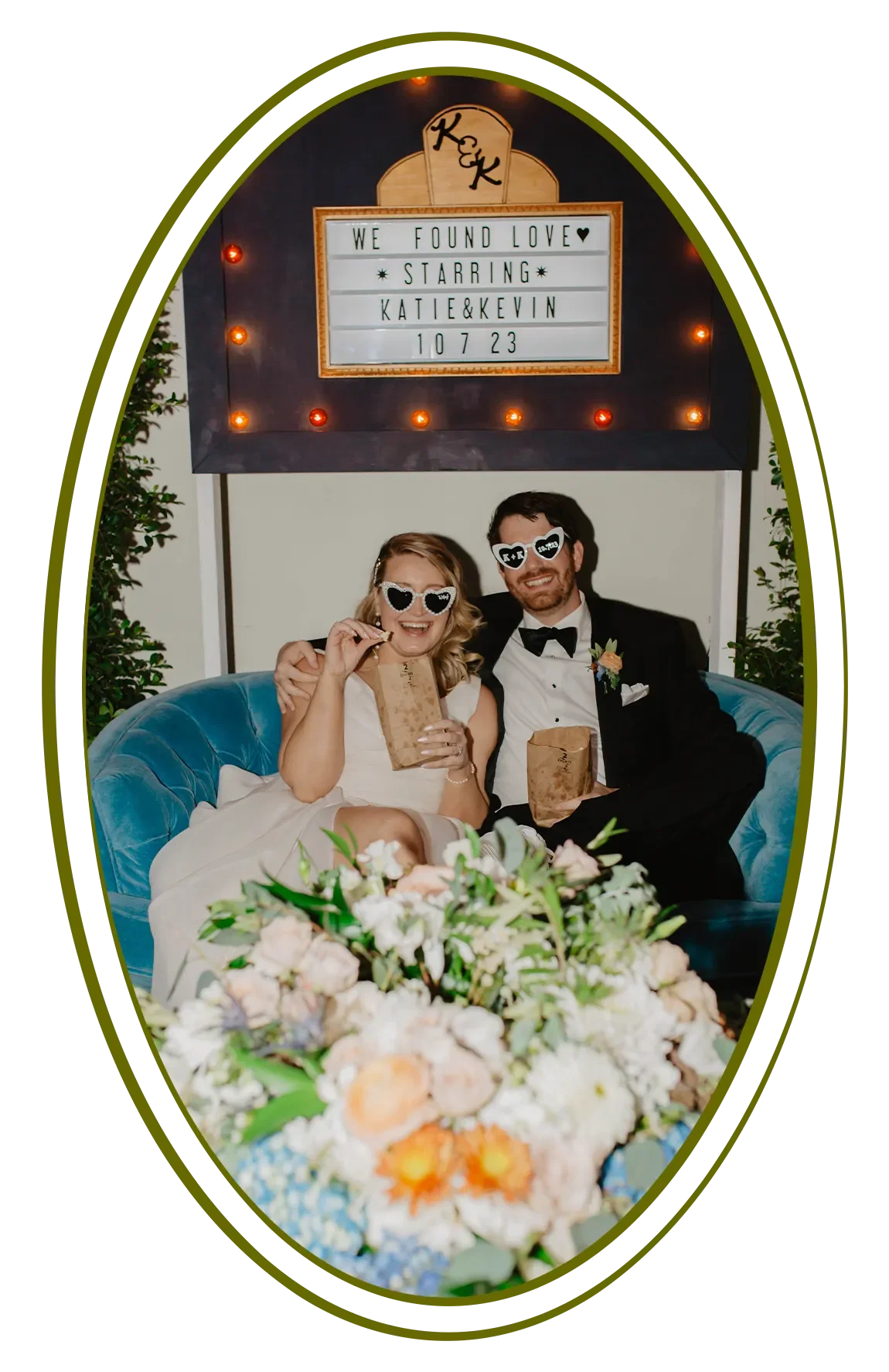 A happy couple in wedding attire wearing heart-shaped sunglasses sitting on a blue couch, holding bags of popcorn. There is a large floral arrangement in front of them, with a backdrop featuring a marquee sign and string lights.