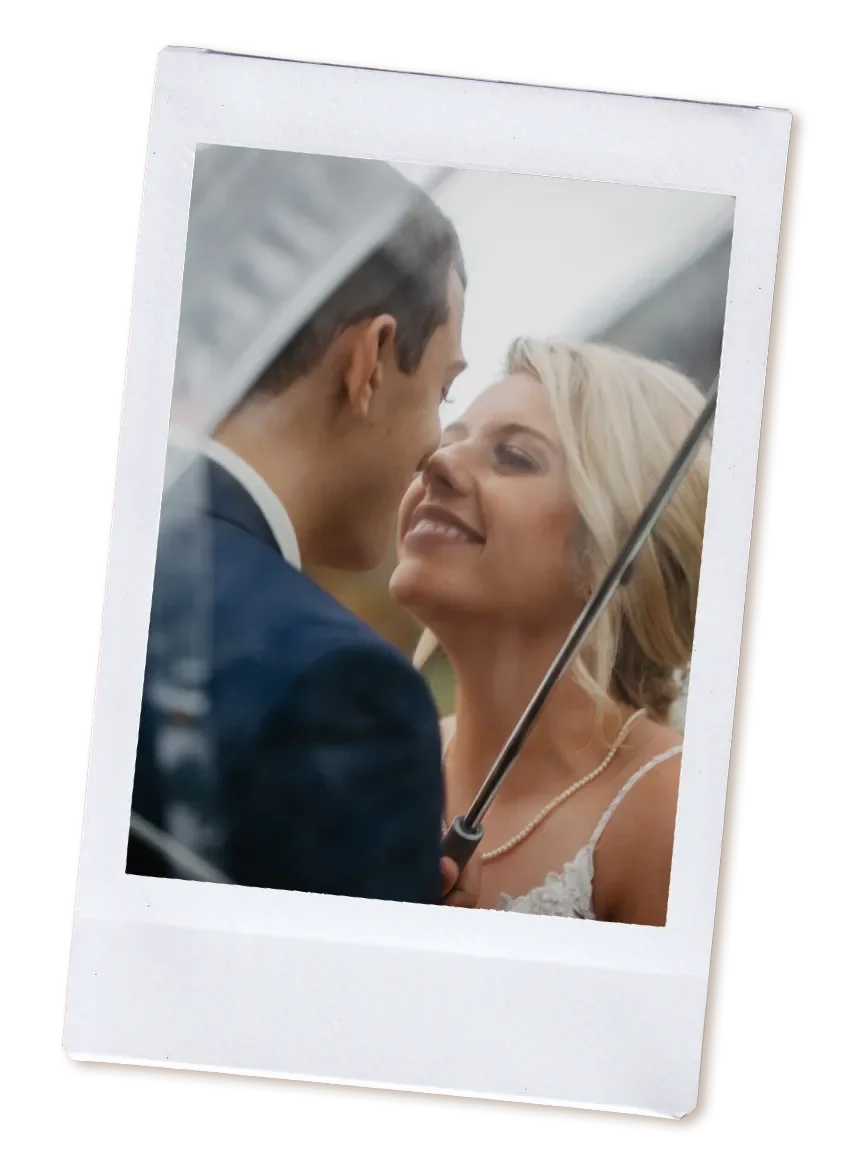 A couple smiling and close together under an umbrella, outdoors, with blurred background.