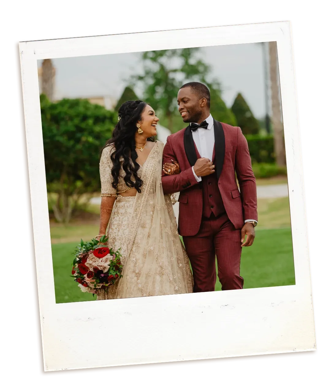A couple dressed in wedding attire, smiling and walking outdoors with a green background. The groom is in a burgundy suit, and the bride is in an intricately beaded beige gown.